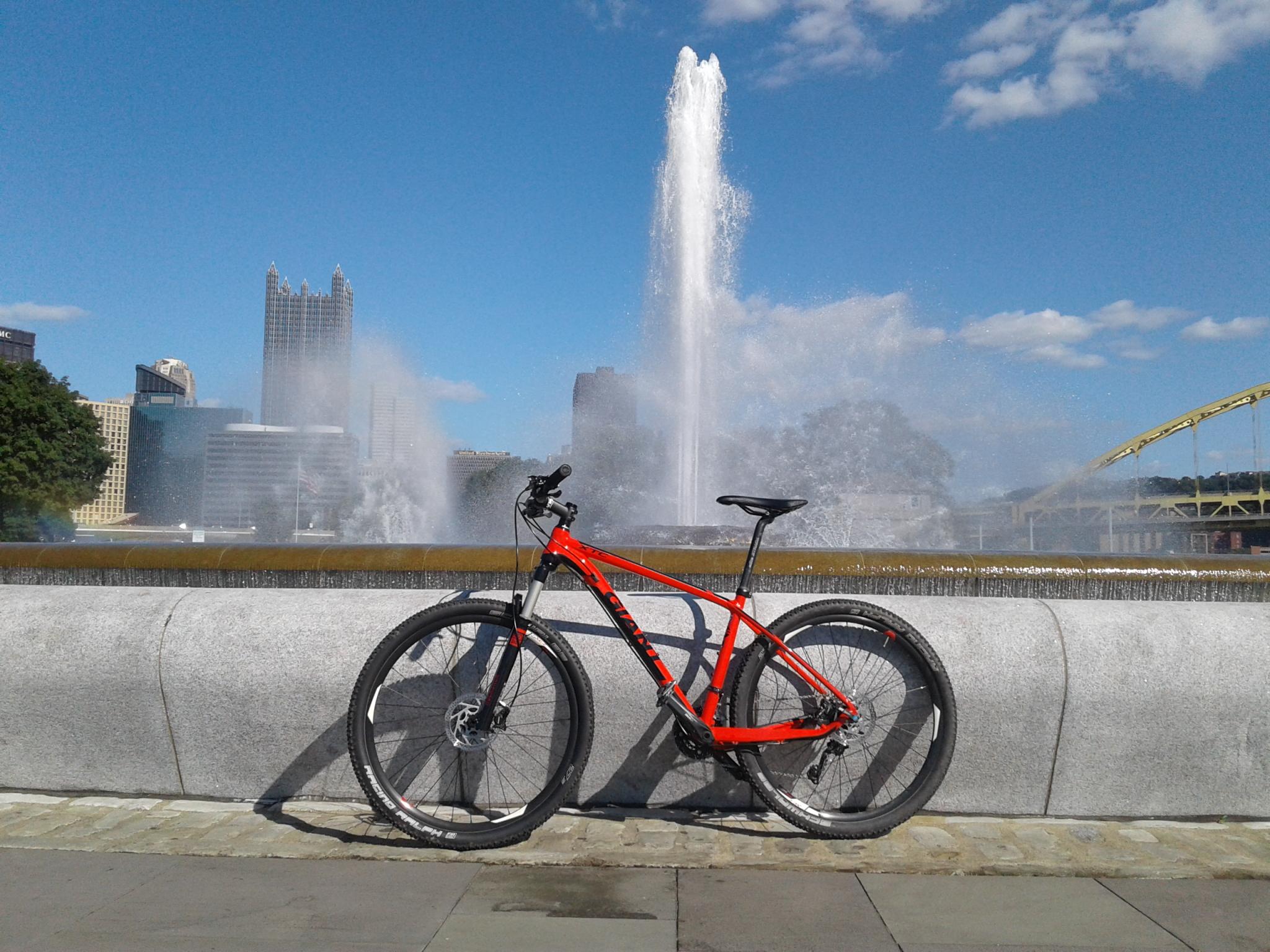 Giant XTC 27.5 2: A bright red mountain bike is leaning against a stone wall near a large fountain, with water spraying upwards. In the background, the skyline of a city can be seen, featuring tall buildings and a bridge. The sky is clear with a few white clouds.