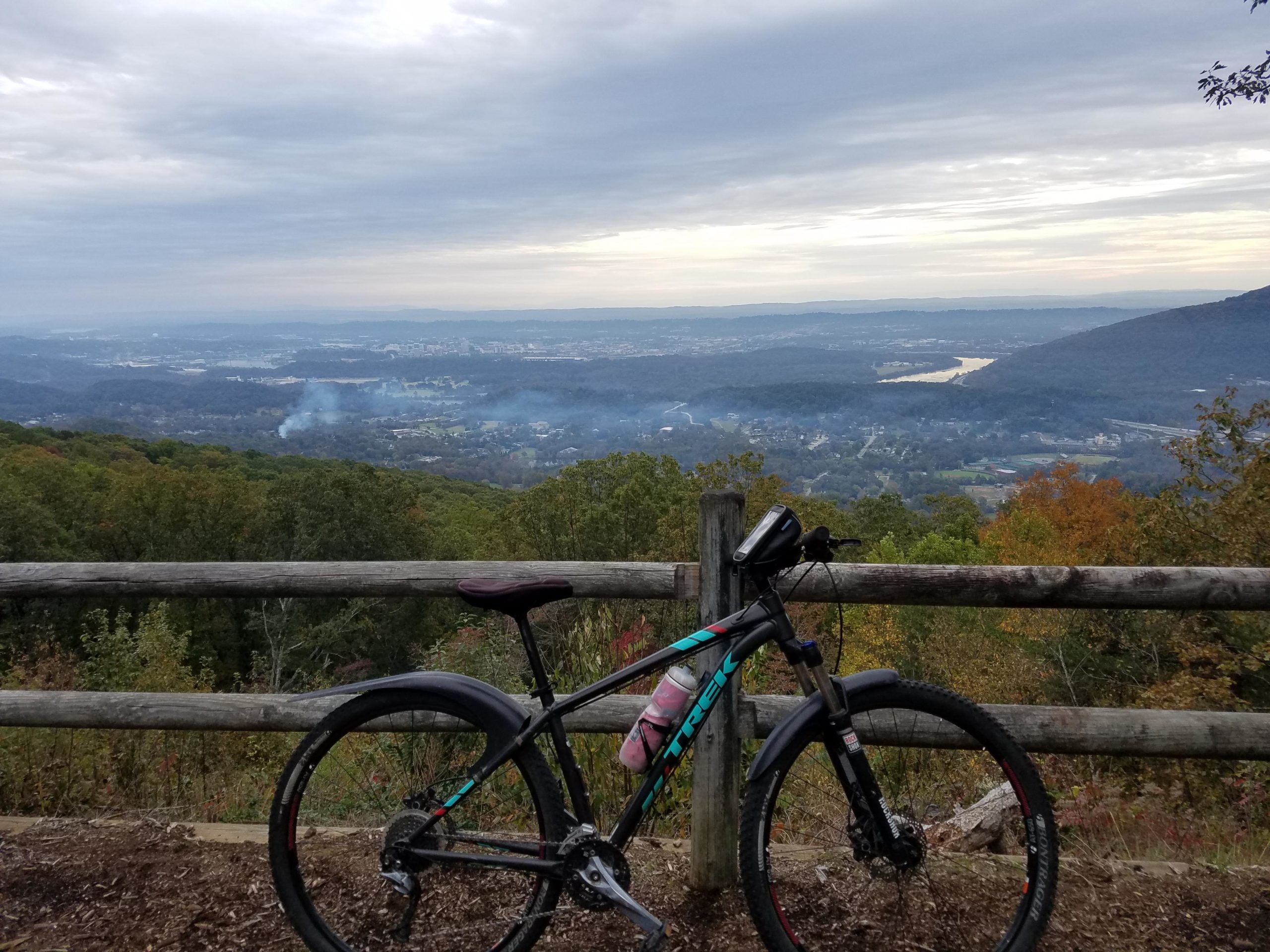 A mountain bike parked beside a wooden fence, overlooking a scenic valley filled with trees and rolling hills under a cloudy sky. Smoke can be seen rising from the landscape in the distance, suggesting nearby activities. The scene captures the beauty of nature and outdoor adventure. East Rim Trail mountain bike trail.