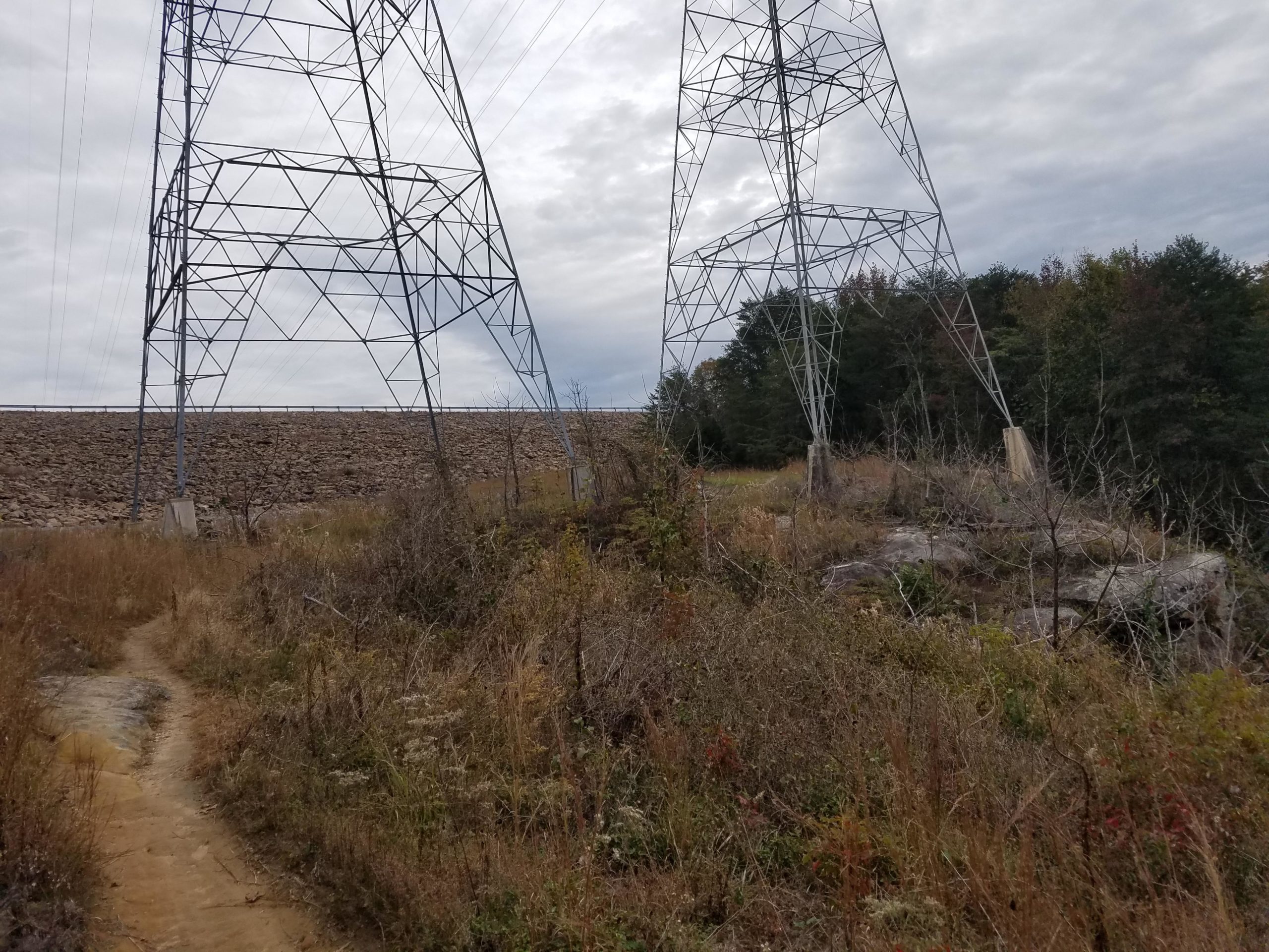 Electricity transmission towers stand near a rocky embankment, surrounded by tall grasses and sparse vegetation. A narrow dirt path winds through the landscape, leading toward the towers under a cloudy sky. Raccoon Mountain Trail Network mountain bike trail.
