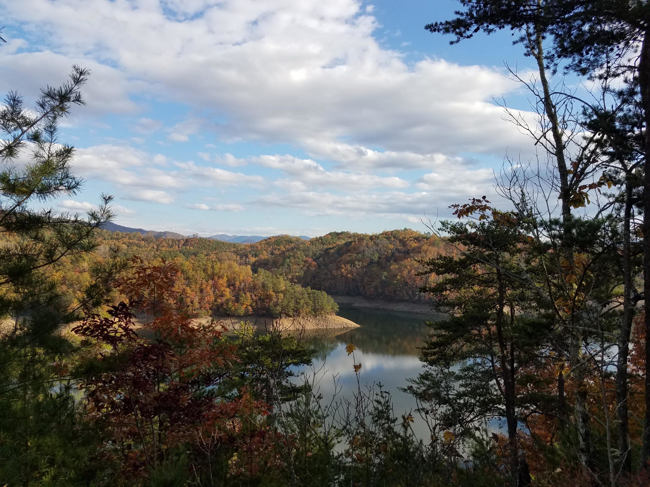 Scenic view of a tranquil lake surrounded by lush, colorful trees in autumn, with rolling hills and a partly cloudy sky above. Tsali Recreation Area mountain bike trail.