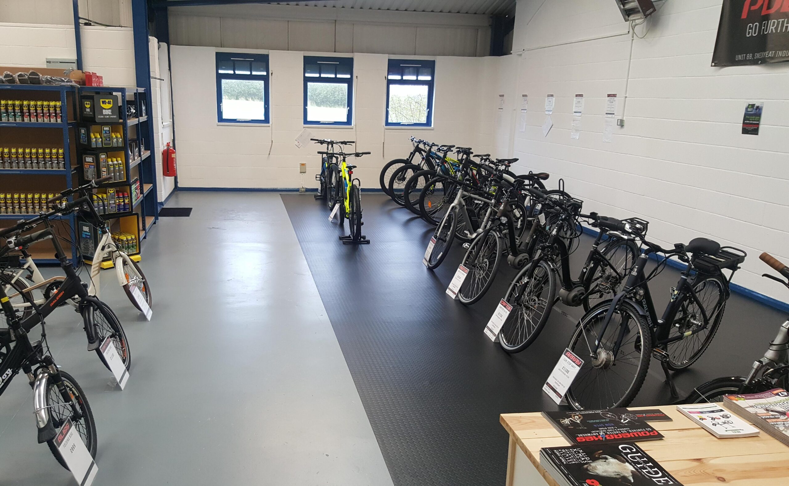 A well-lit bike showroom featuring a row of various bicycles for sale, displayed on a rubber floor. On the left, a shelf stocked with bike maintenance products. The walls are white, and there are several windows allowing natural light into the space. Promotional materials and price tags are visible on the bicycles.