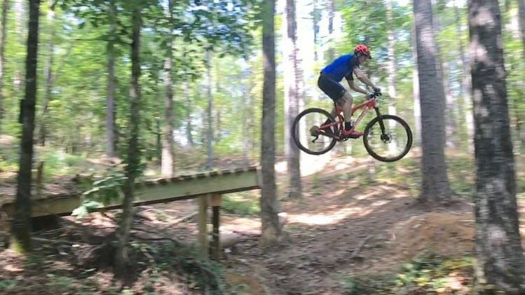 Santa Cruz Tallboy Carbon: A mountain biker in a blue shirt and red helmet jumps off a wooden bridge in a forested area, surrounded by green trees and underbrush.