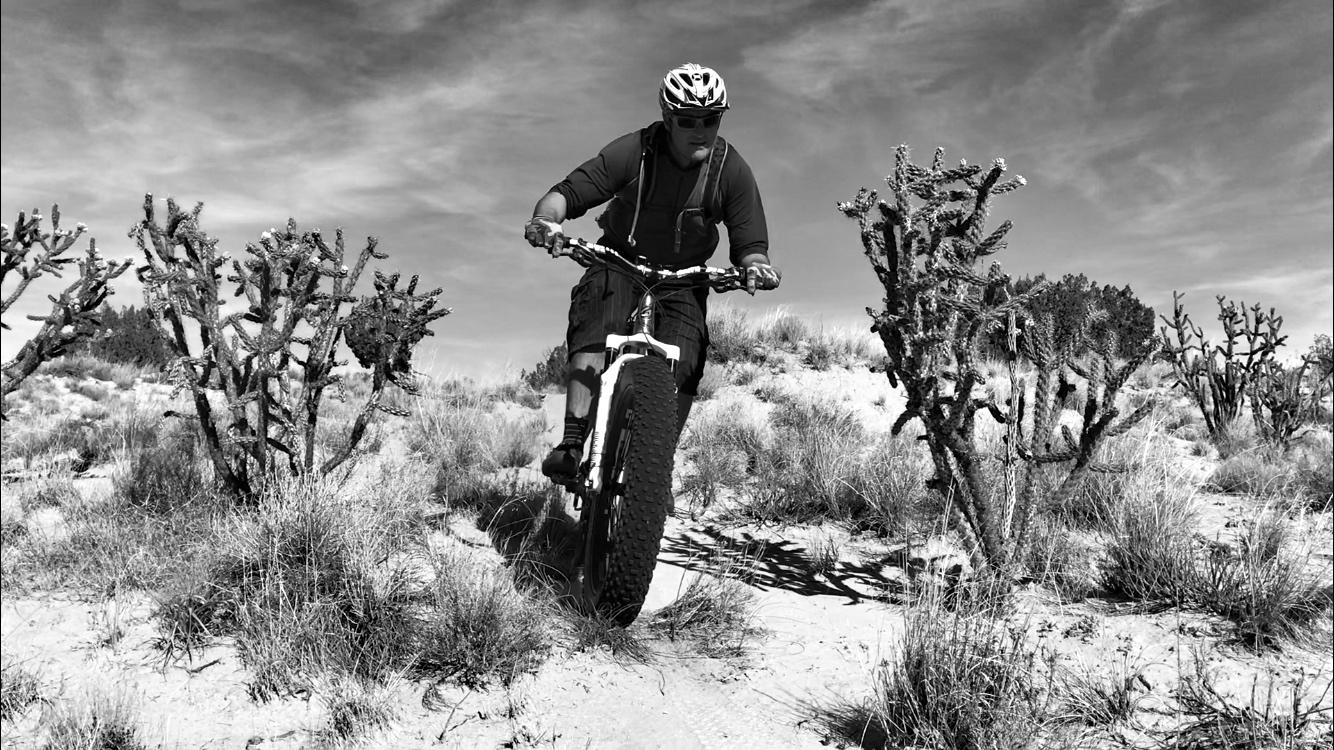 A person riding a mountain bike on a sandy trail surrounded by cacti and desert vegetation, captured in black and white. The cyclist is focused, positioned over the bike as they navigate the terrain under a cloudy sky. Mariposa Fat Bike Trails mountain bike trail.