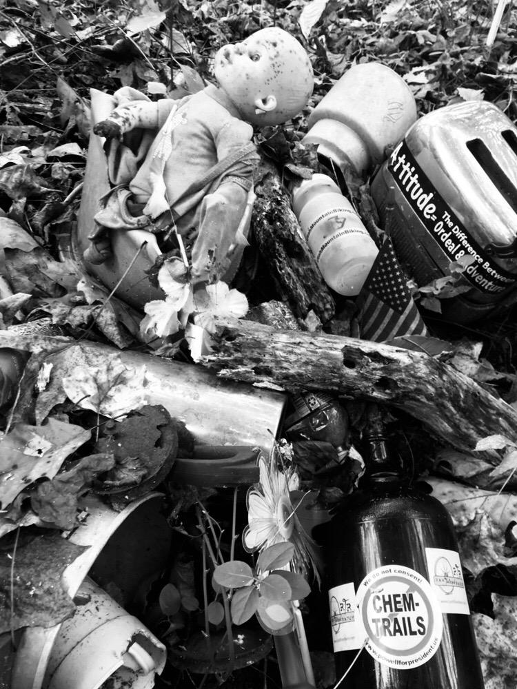 A black and white image depicting a cluttered scene of discarded items, including a dirty doll, various plastic bottles, a helmet, and a black bottle labeled "CHEM-TRAILS." The ground is covered with fallen leaves and debris, creating a sense of neglect and environmental pollution. Rosaryville State Park mountain bike trail.