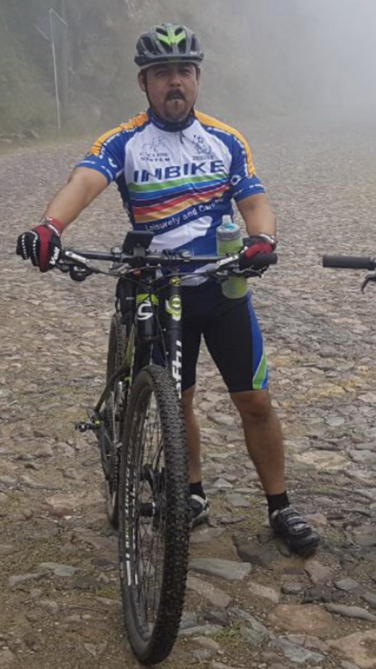 Cannondale F29 6: A cyclist in a colorful jersey stands next to a mountain bike on a rocky path. The cyclist has a serious expression and is wearing black gloves and cycling shoes. The background features a foggy atmosphere, suggesting a mountainous or rural setting.