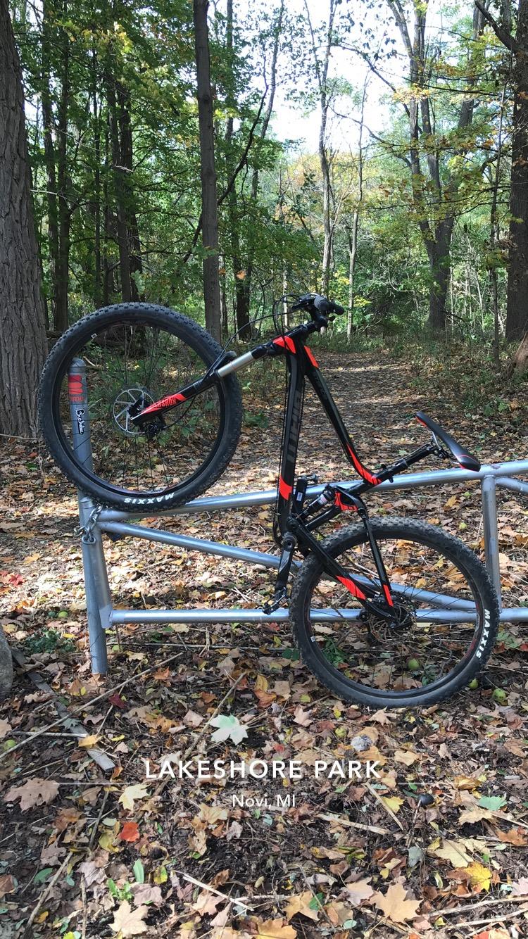 Giant Stance 27.5 2: A black and red mountain bike resting on a silver railing in Lakeshore Park, surrounded by autumn foliage and trees. The ground is covered with fallen leaves, and a trail is visible in the background.