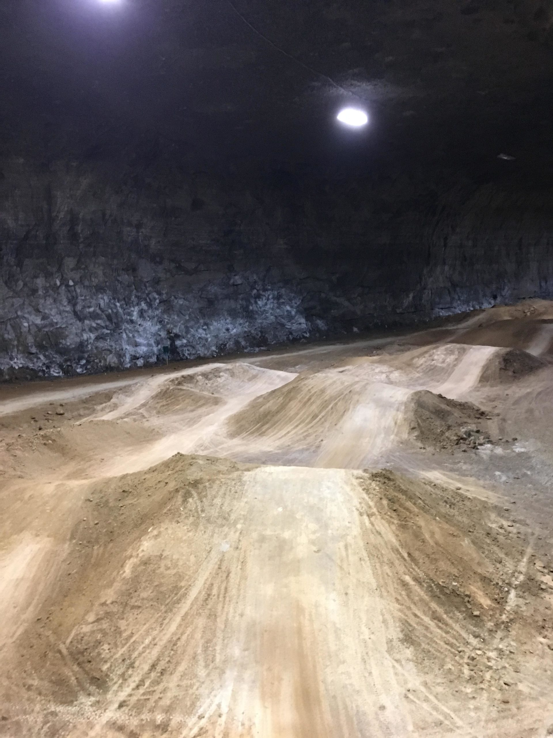 A wide, underground track featuring mounds of dirt and a smooth, winding path, illuminated by overhead lights against a rocky background. The scene suggests an indoor biking or motocross course with a rugged terrain. Louisville Mega Cavern Bike Park mountain bike trail.