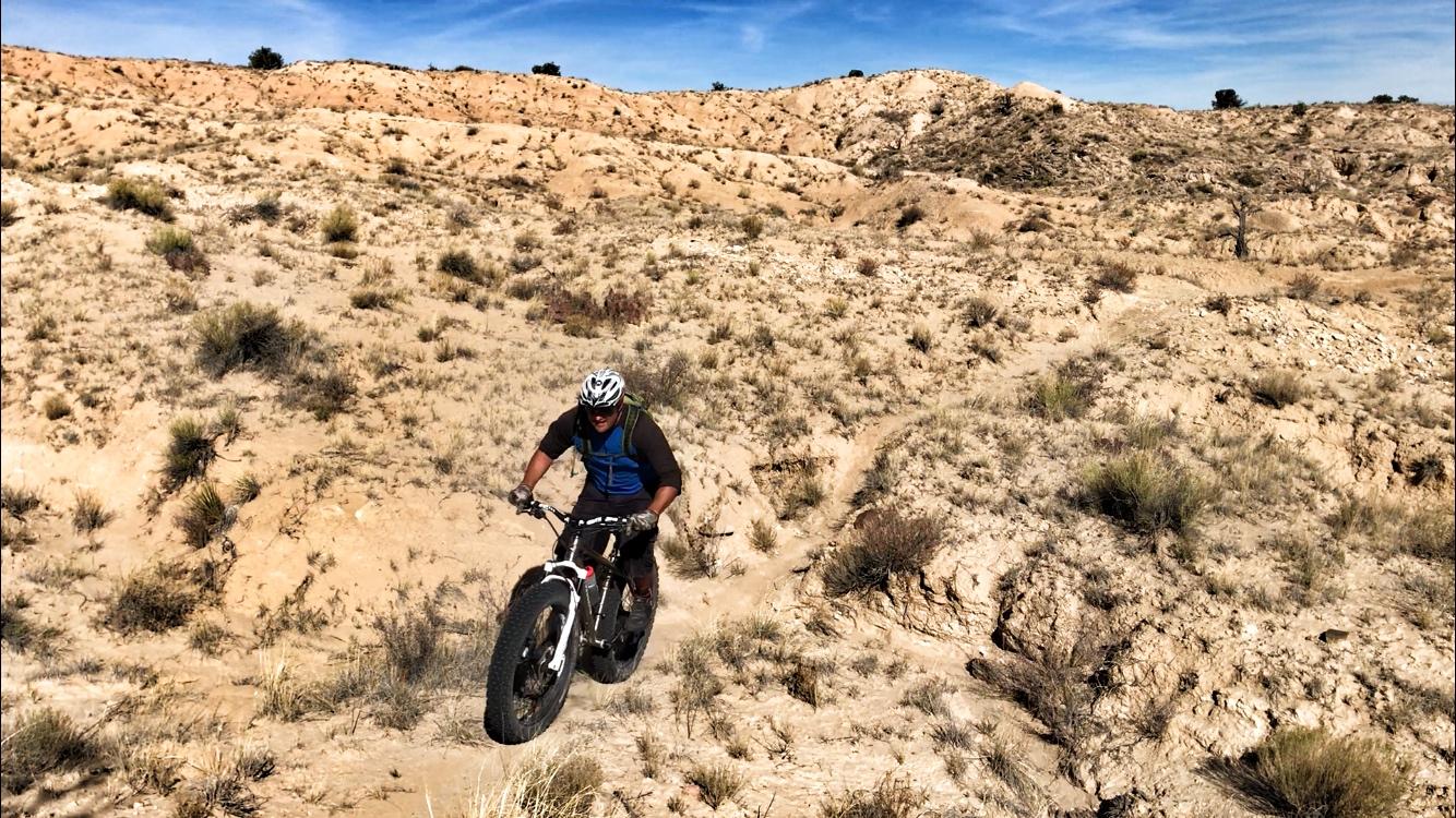 A mountain biker ascends a rocky, arid terrain with sparse vegetation and dry hills in the background, under a clear blue sky. The biker is wearing a helmet and a blue and brown outfit, navigating the uneven path on a fat tire bike. Mariposa Fat Bike Trails mountain bike trail.