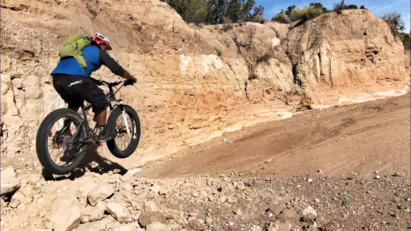 A mountain biker performing a jump over rocky terrain, with a steep, sandy cliff in the background and sparse vegetation. The rider is wearing a helmet, a blue shirt, and black shorts, with a green backpack. Mariposa Fat Bike Trails mountain bike trail.