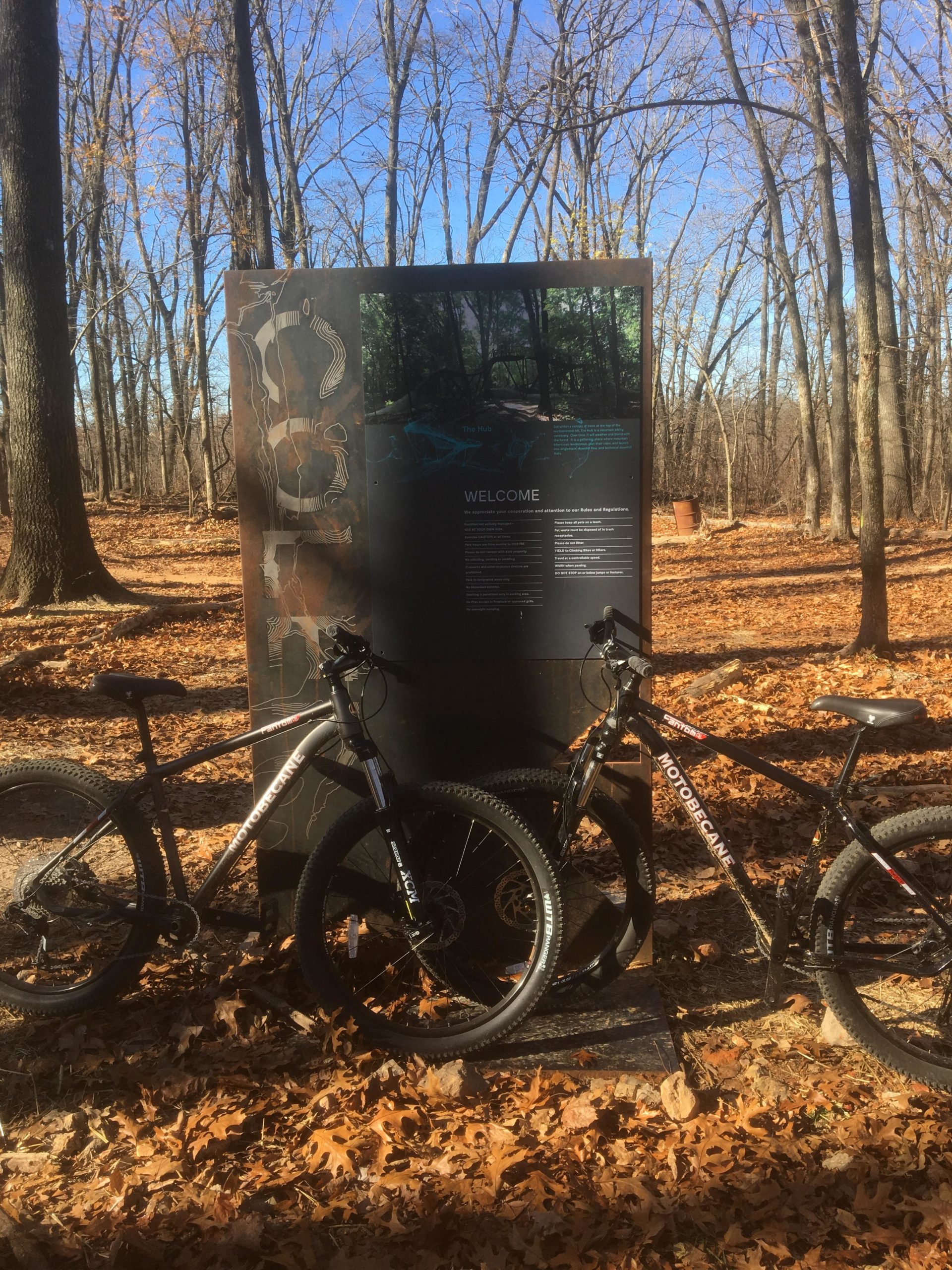 Two mountain bikes parked beside a trail information sign in a wooded area with autumn leaves covering the ground. The sign provides details about the trail, including a welcome message and a map layout, against a backdrop of bare trees and a clear blue sky. Coler Mountain Bike Preserve mountain bike trail.