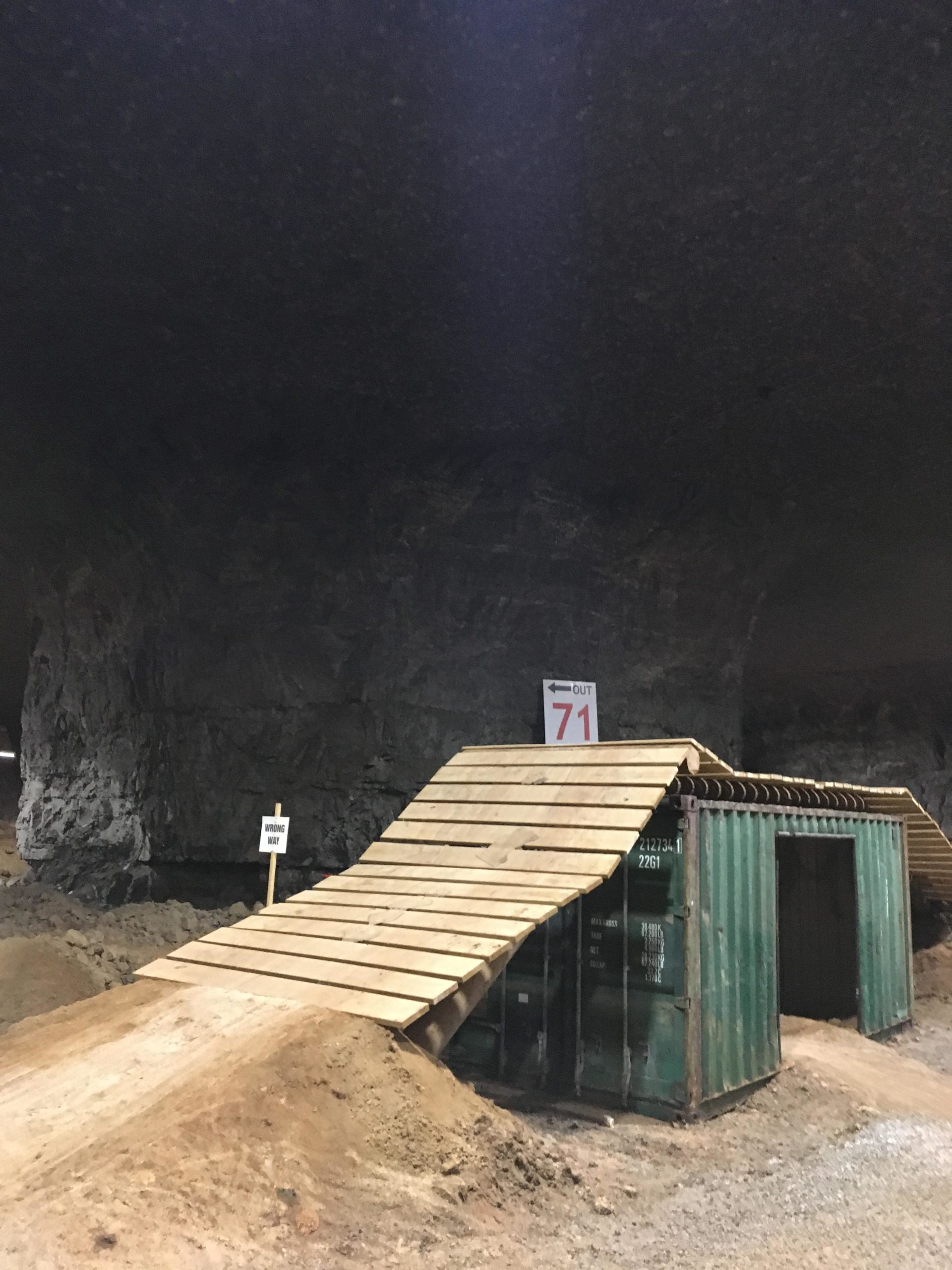 A green shipping container situated in a cavernous, underground space, featuring a wooden ramp leading to it. The ramp is surrounded by loose dirt and gravel, with a sign indicating "OUT 71" nearby. The ceiling of the cavern is dark and rocky, reflecting an industrial environment. Louisville Mega Cavern Bike Park mountain bike trail.