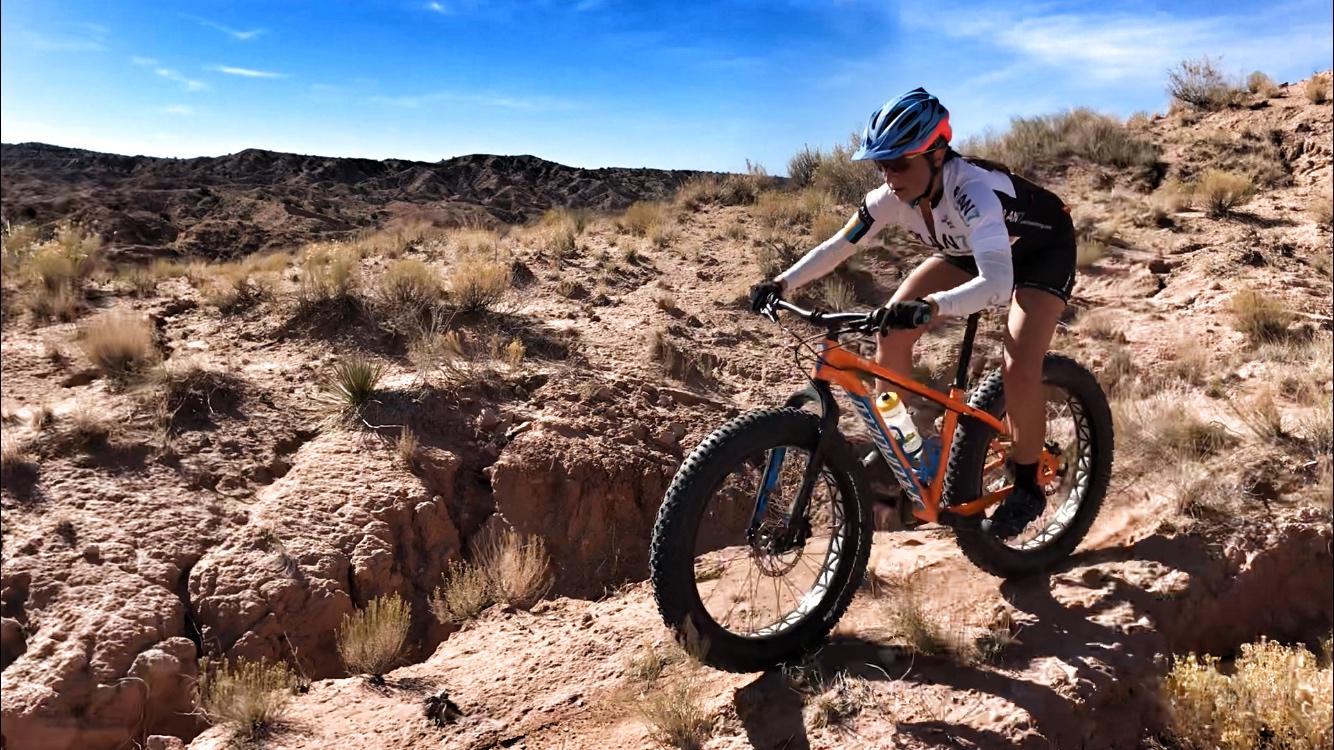 A person riding a mountain bike on a rocky trail in a desert landscape. The cyclist is wearing a helmet and athletic gear, navigating a challenging section of the path surrounded by sparse vegetation and rugged terrain under a clear blue sky. Mariposa Fat Bike Trails mountain bike trail.