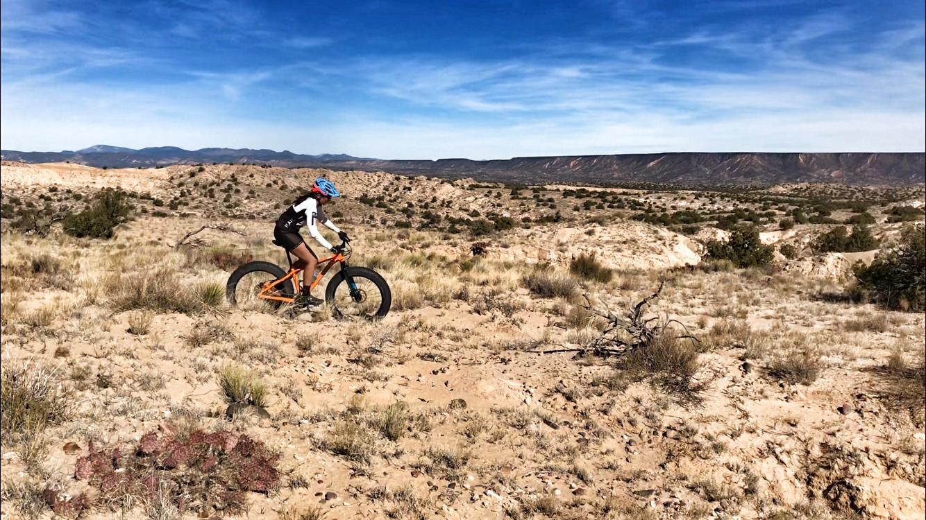 A cyclist wearing a blue helmet and a black and white jersey rides a bright orange fat bike across a rocky, desert landscape with sparse vegetation and rolling hills under a clear blue sky. Mariposa Fat Bike Trails mountain bike trail.
