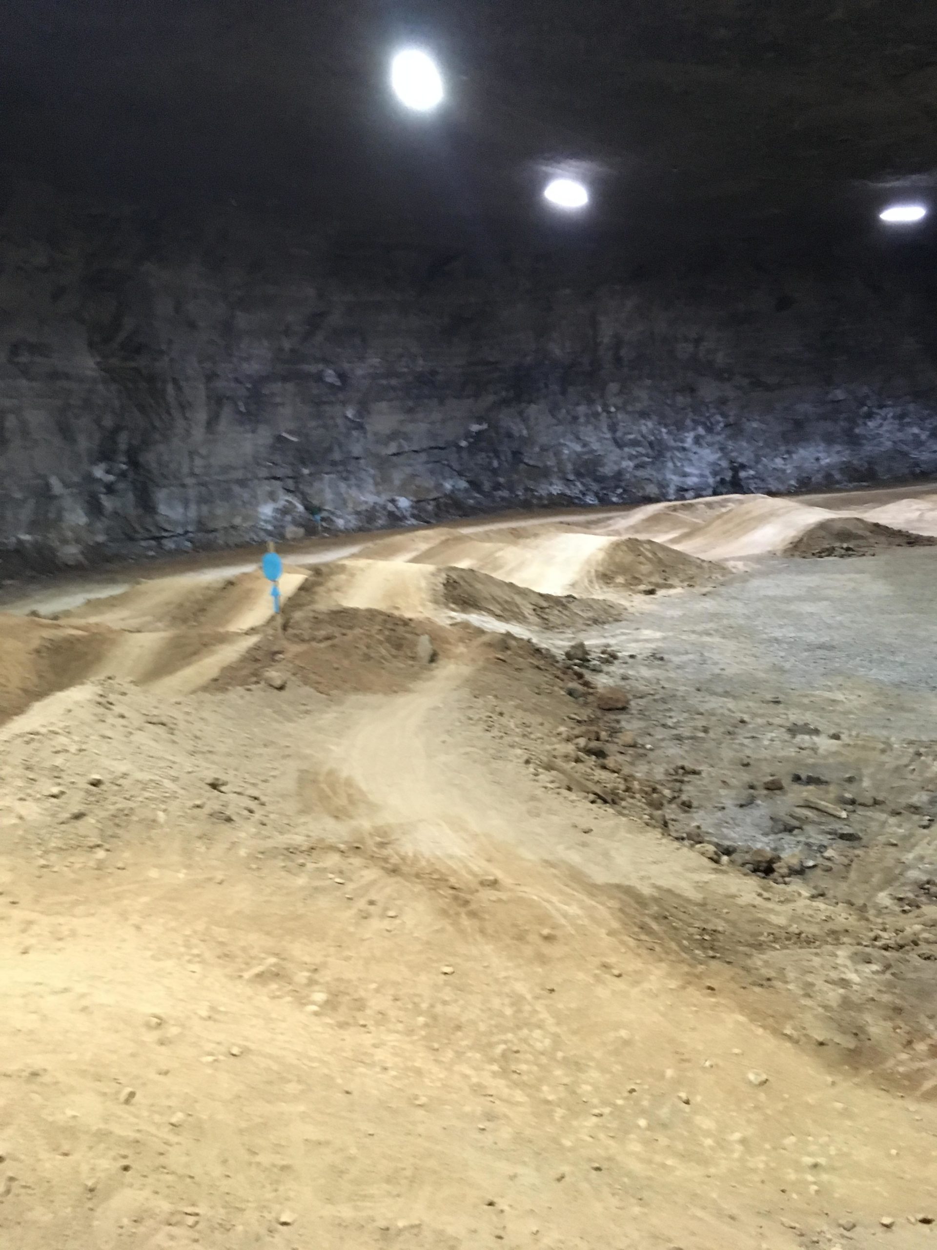Alt text: An interior view of an expansive underground space with dirt paths and mounds, illuminated by circular lights in the ceiling. The rocky walls are visible, providing a natural backdrop to the dirt terrain. Louisville Mega Cavern Bike Park mountain bike trail.