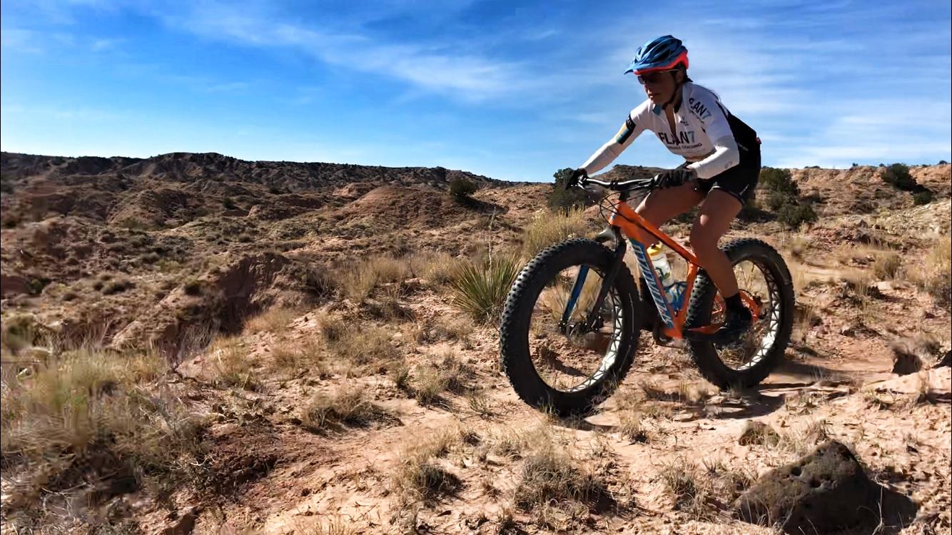 A person riding a mountain bike on a rugged terrain under a blue sky. The cyclist is dressed in a jersey and biking shorts, wearing a helmet, and navigating through a landscape of hills and sparse vegetation. The scene captures the excitement of outdoor biking in a natural setting. Mariposa Fat Bike Trails mountain bike trail.