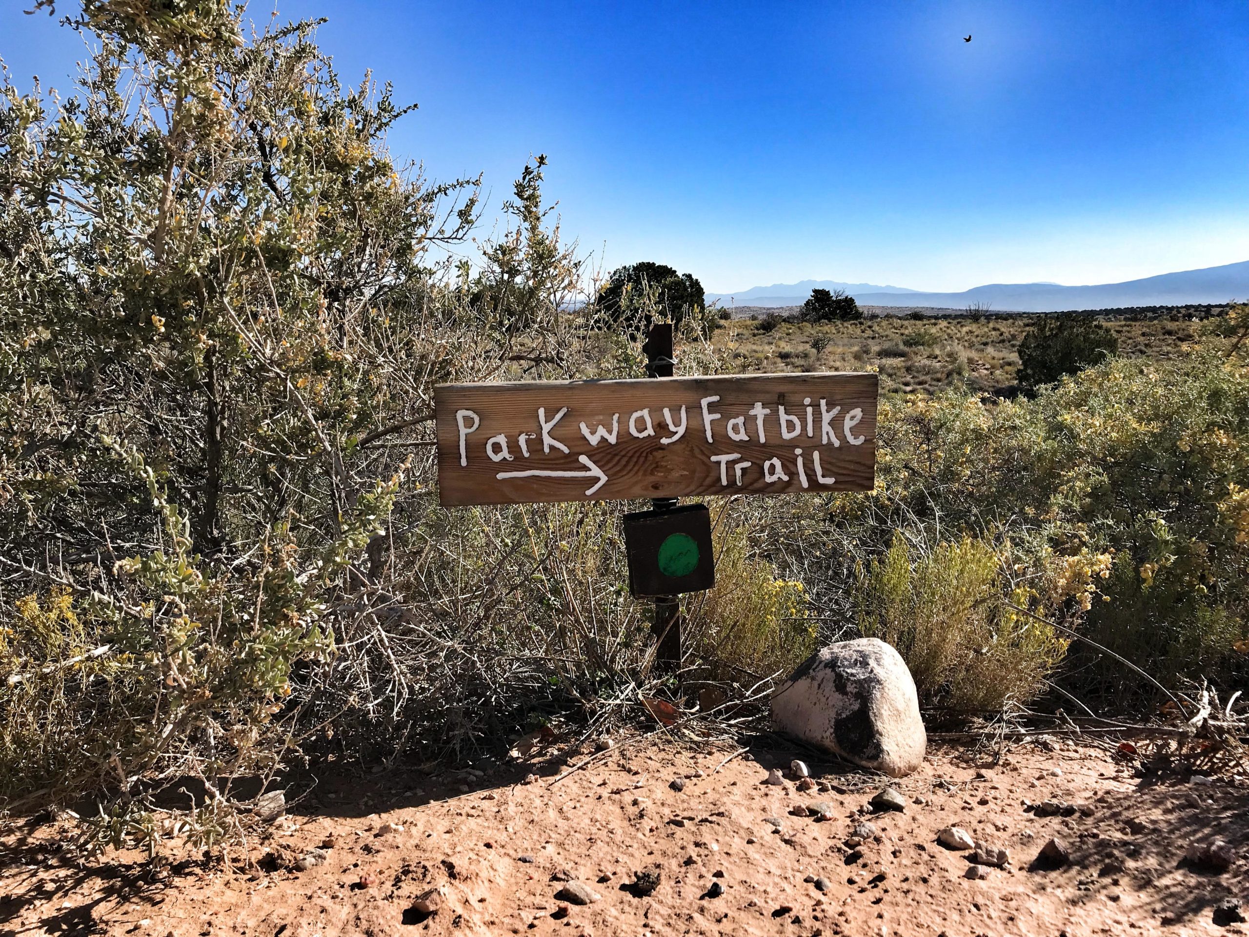 Wooden sign indicating the direction to the "Parkway Fatbike Trail," surrounded by desert vegetation and a clear blue sky. The trail sign points left, with a green circular marker below it. Parkway Fatbike trail mountain bike trail.