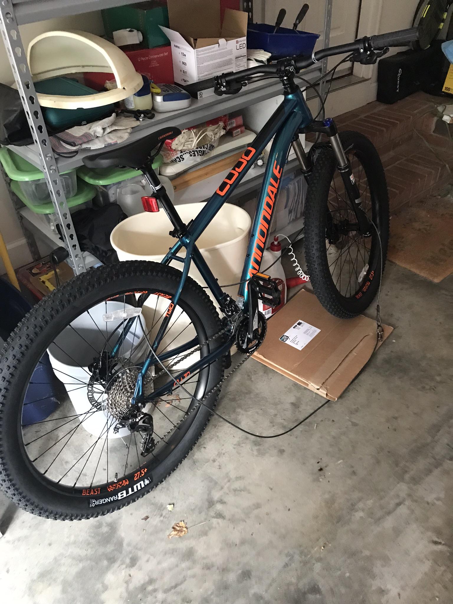 Cannondale Cujo 2: A blue Cannondale mountain bike with orange accents, parked in a cluttered garage. Various storage bins and items are visible in the background, along with a cardboard box on the ground beneath the bike. The bike features wide tires designed for off-road use.
