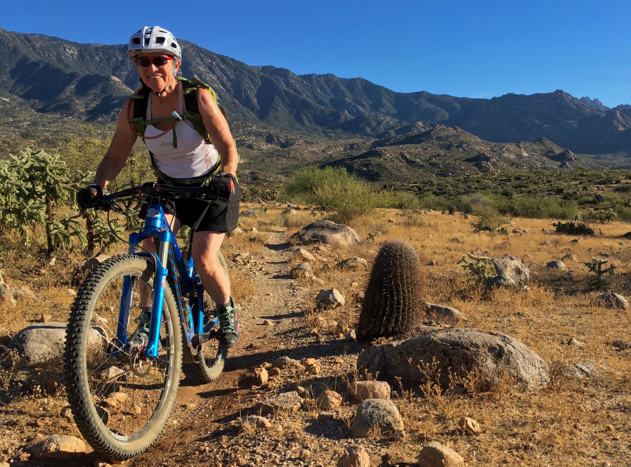 A woman rides a blue mountain bike along a rocky trail in a desert landscape, with mountains in the background and cacti scattered around. She is wearing a helmet and sunglasses, dressed in a tank top and shorts, enjoying the sunny day. 50-year Trail / Golder Ranch mountain bike trail.