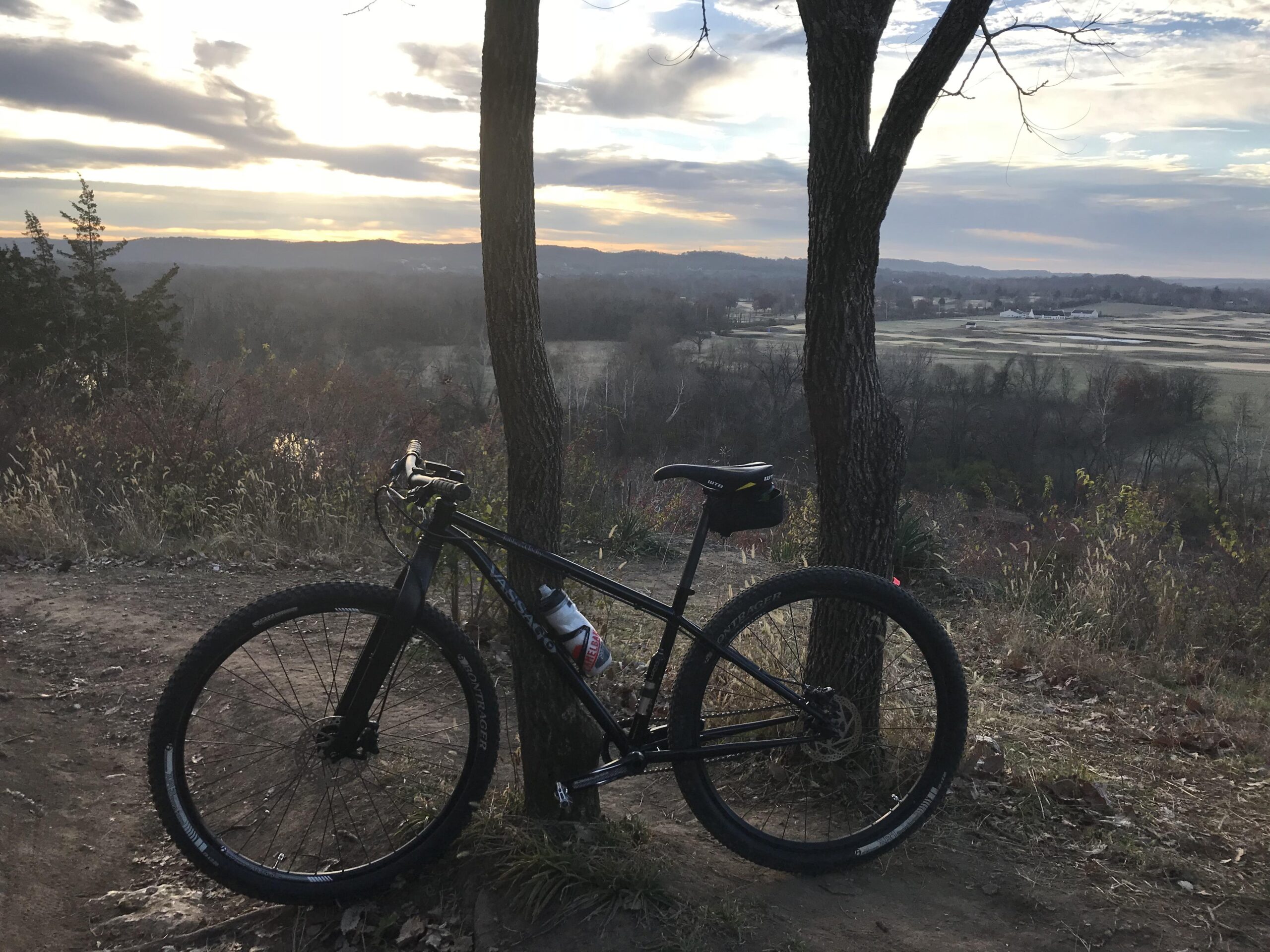 Vassago Jabberwocky: A mountain bike resting against a tree along a scenic trail, with a panoramic view of rolling hills and fields in the background under a partly cloudy sky.