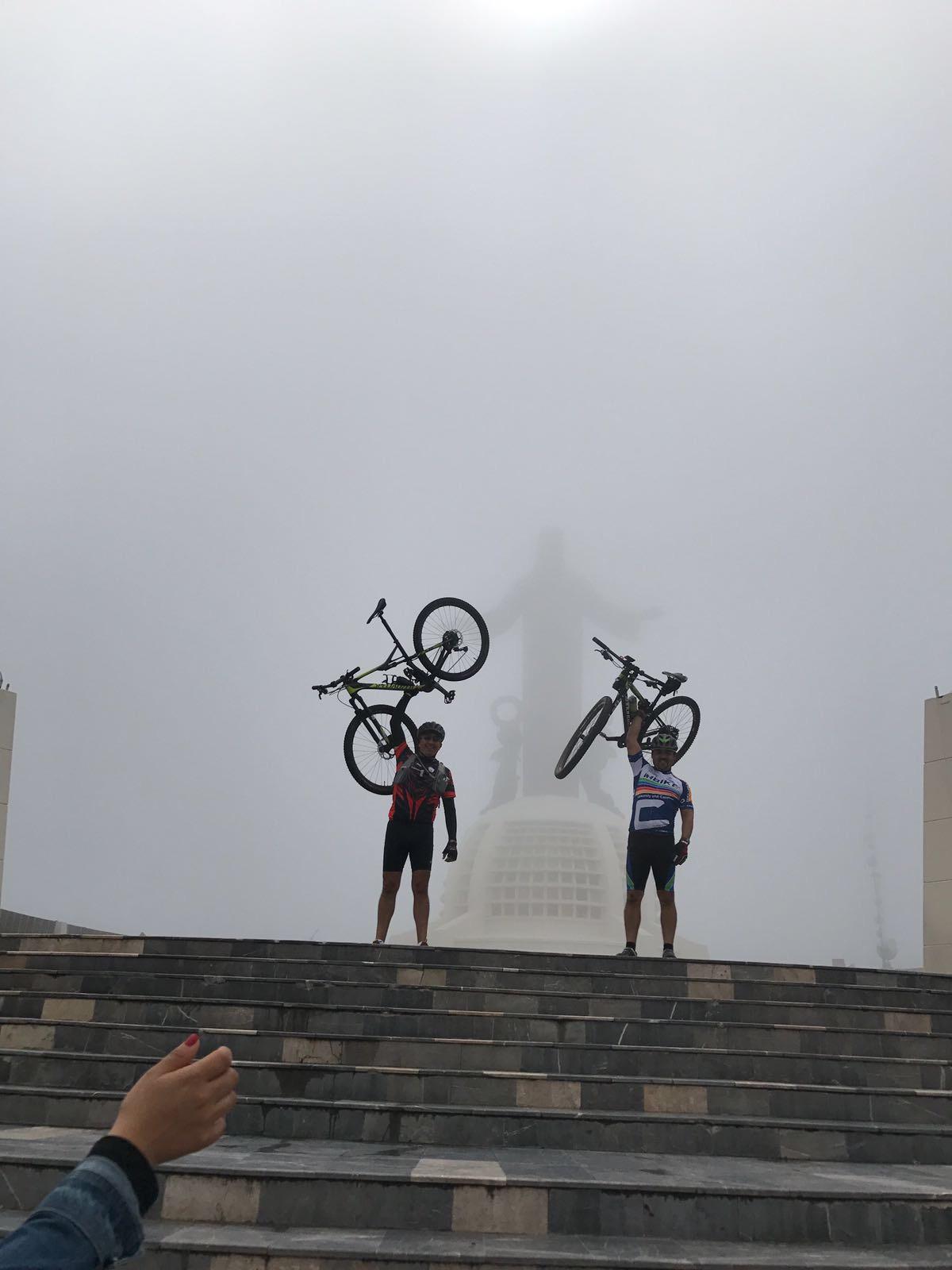 Cannondale F29 6: Two cyclists standing on a staircase, raising their bicycles above their heads in celebration. The background is shrouded in fog, obscuring the details of their surroundings, while a large monument is faintly visible through the mist. One cyclist is wearing a red and black outfit, while the other is dressed in blue and white. A hand can be seen in the lower corner of the image.