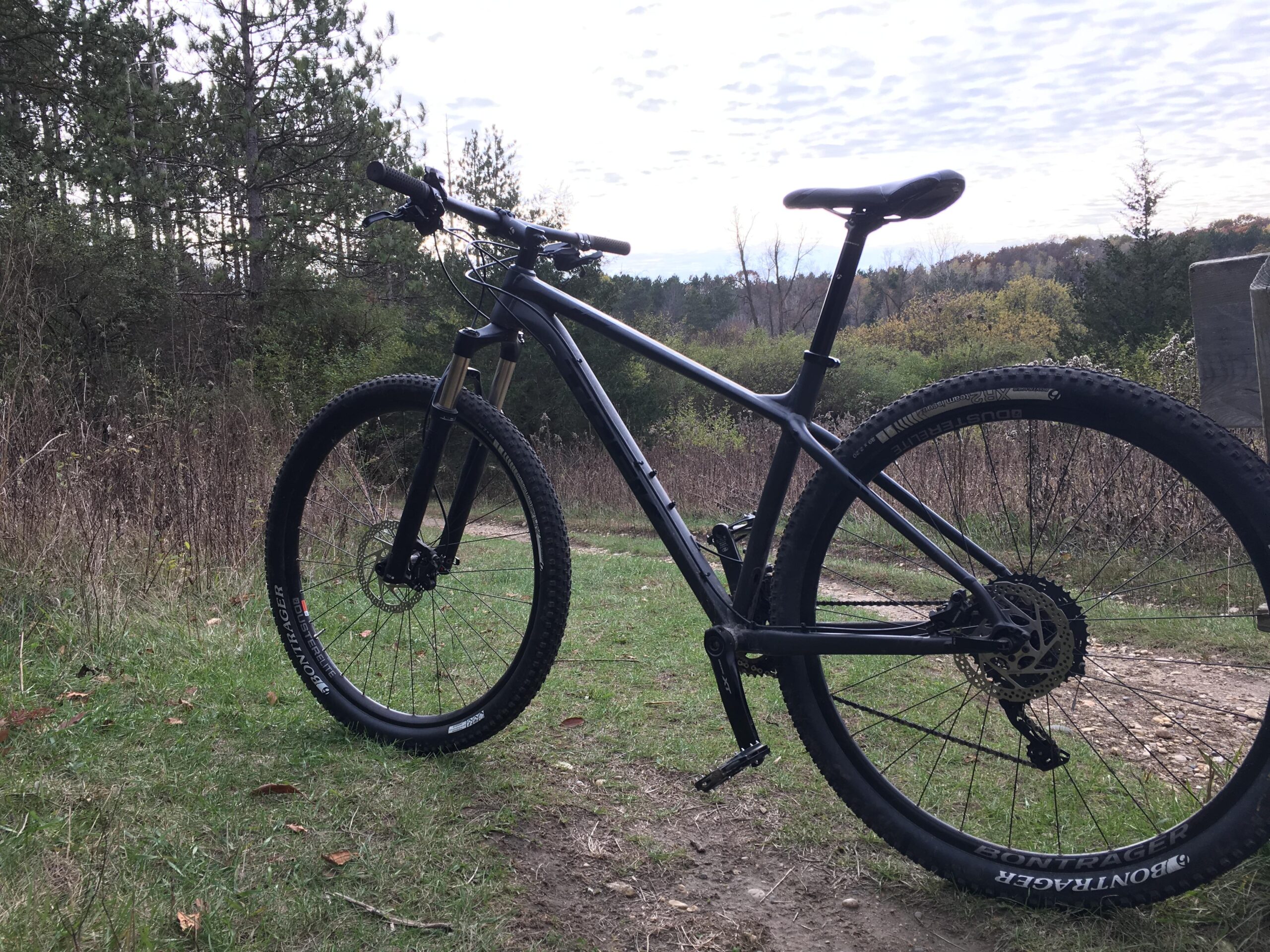 Trek X Caliber 9: A mountain bike positioned on a grassy trail, surrounded by trees and shrubs, with a cloudy sky overhead. The bike features thick tires designed for off-road terrain and a sleek, black frame.
