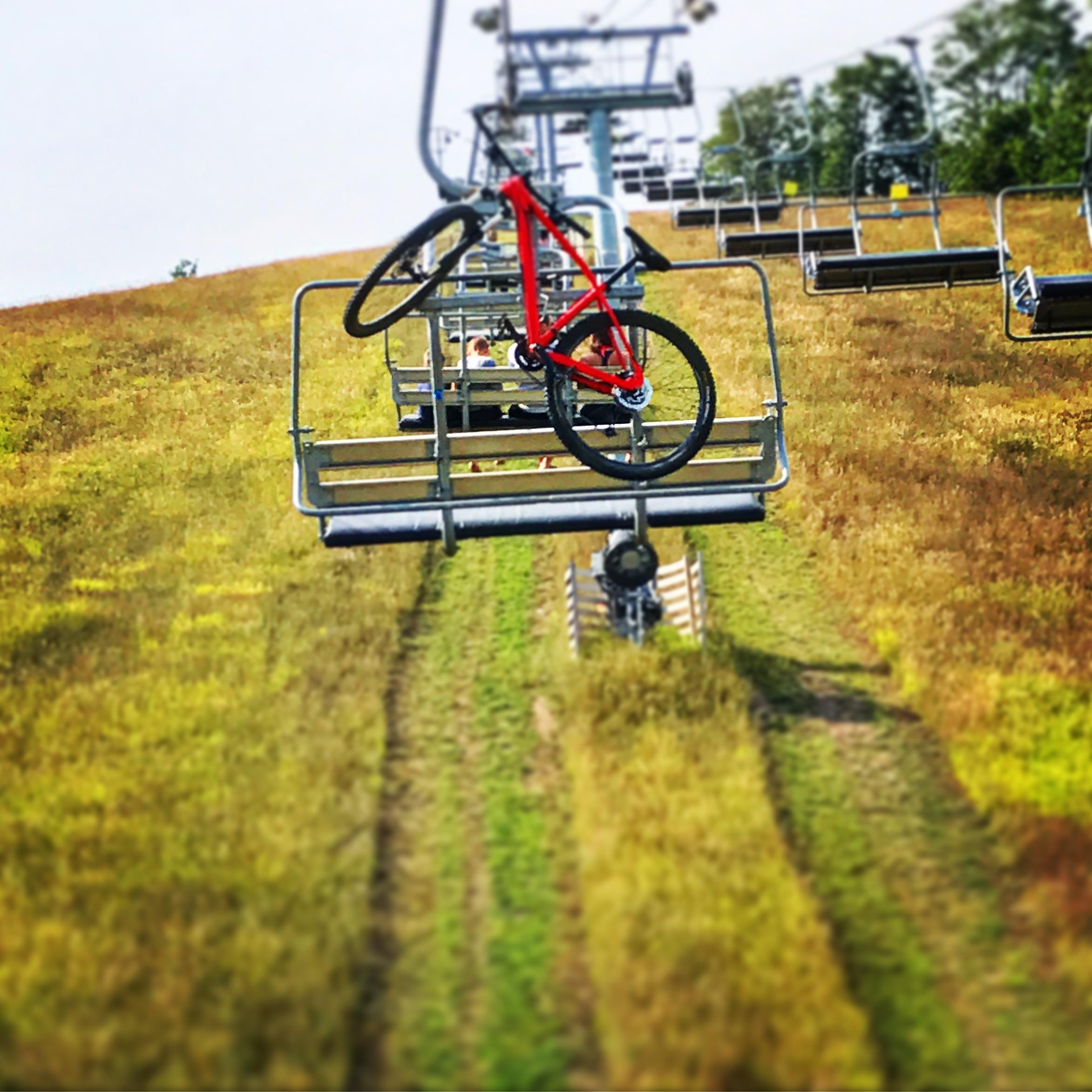 Specialized Rockhopper Comp 29: A red mountain bike is suspended from a ski lift against a backdrop of a grassy hill. The lift chairs are empty, and patches of yellow and green grass cover the slope below.