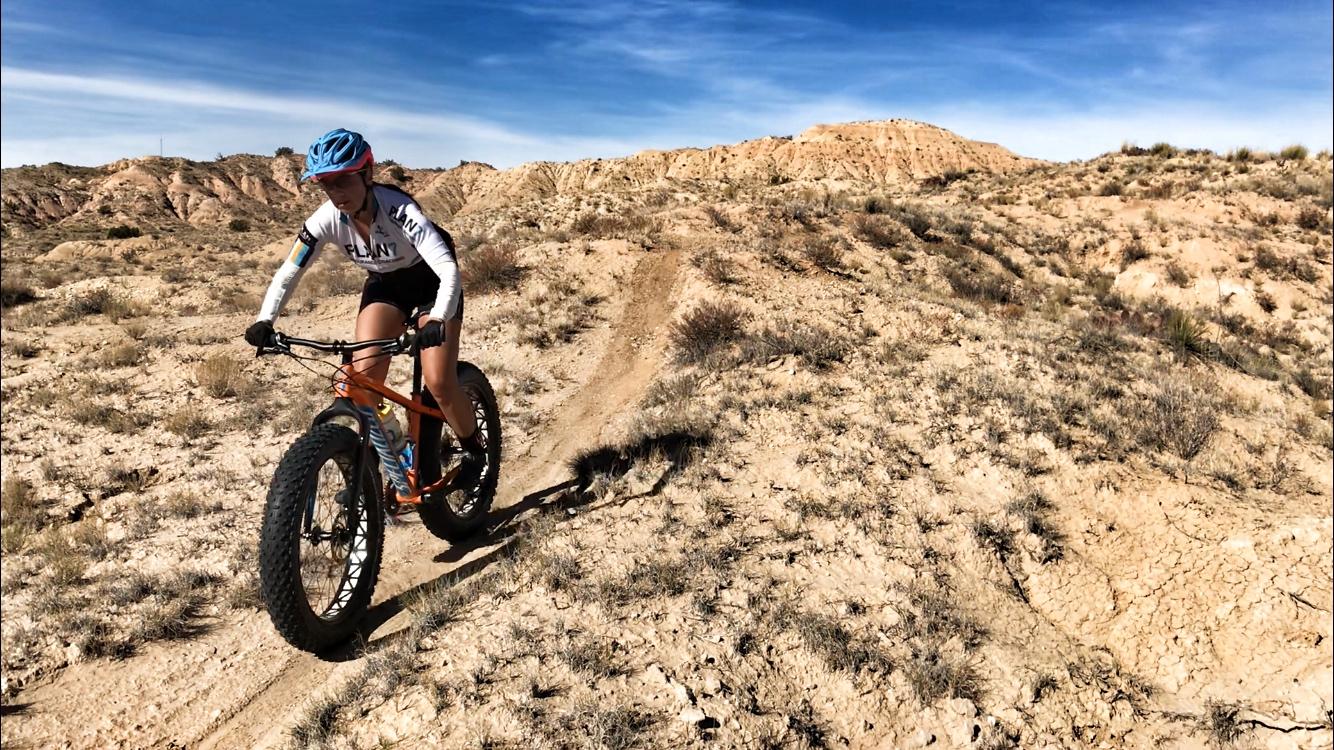 A person riding a fat tire bike on a dirt trail in a rugged desert landscape with hills in the background and blue skies above. The rider is wearing a helmet and athletic gear, navigating through sparse vegetation and rocky terrain. Mariposa Fat Bike Trails mountain bike trail.