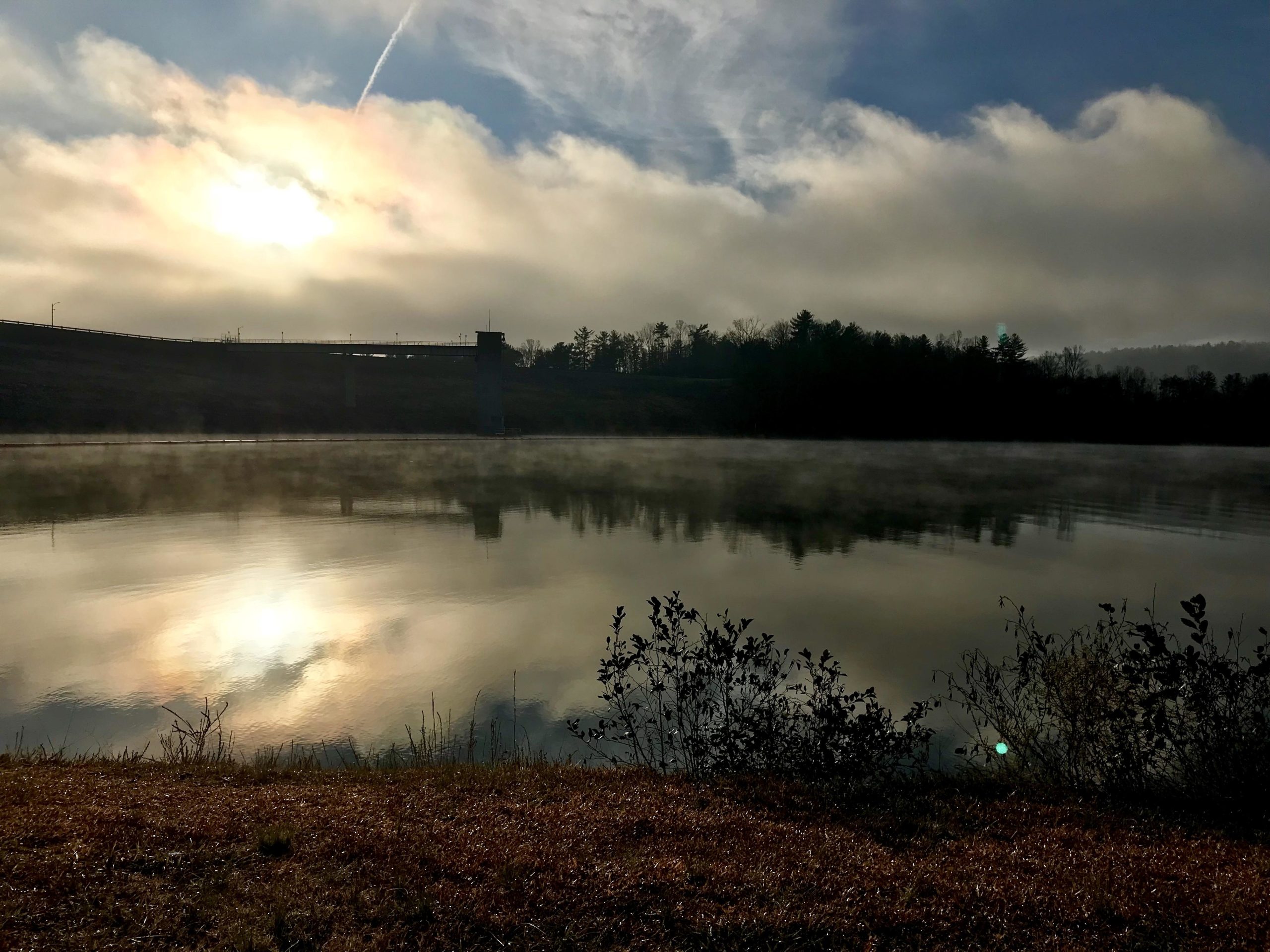 A serene landscape featuring a calm lake reflecting the sun partially hidden by clouds. Mist hovers over the water, with a grassy shoreline and silhouettes of trees in the background. A bridge is visible on the left side, under a light blue sky with wispy clouds. Dark Mountain Trail mountain bike trail.