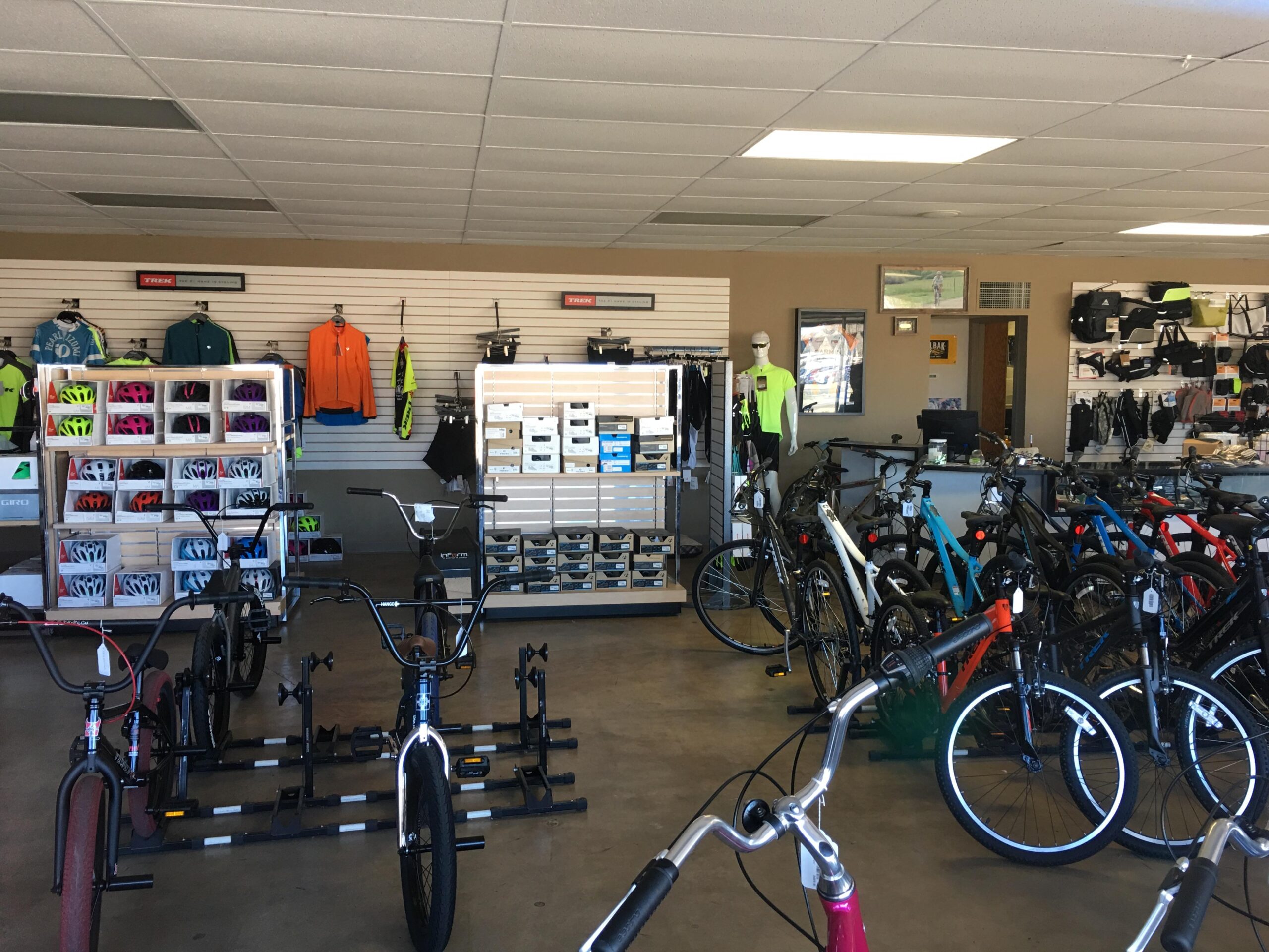 Interior of a bike shop showcasing a variety of bicycles and cycling gear. Shelves are stocked with helmets, clothing, and accessories, while several bikes are displayed prominently on the floor. A mannequin wearing cycling attire stands amidst the merchandise, and a counter is visible in the background.