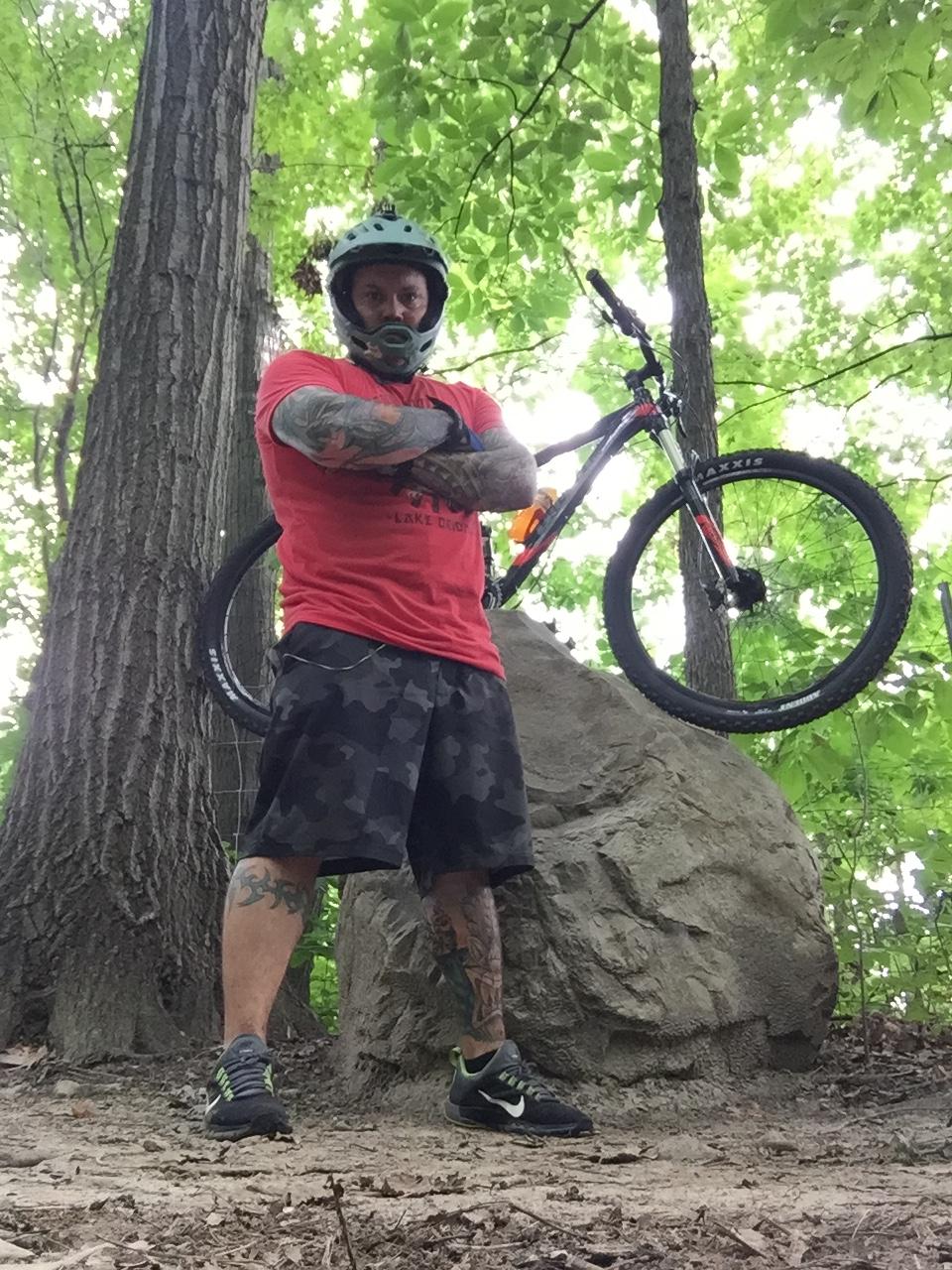 Giant Stance 27.5 2: A person wearing a helmet and a red shirt stands confidently with arms crossed in a forested area. They are on a rocky surface next to a mountain bike, which is resting against a tree. The background features lush green leaves, indicating a natural outdoor setting.
