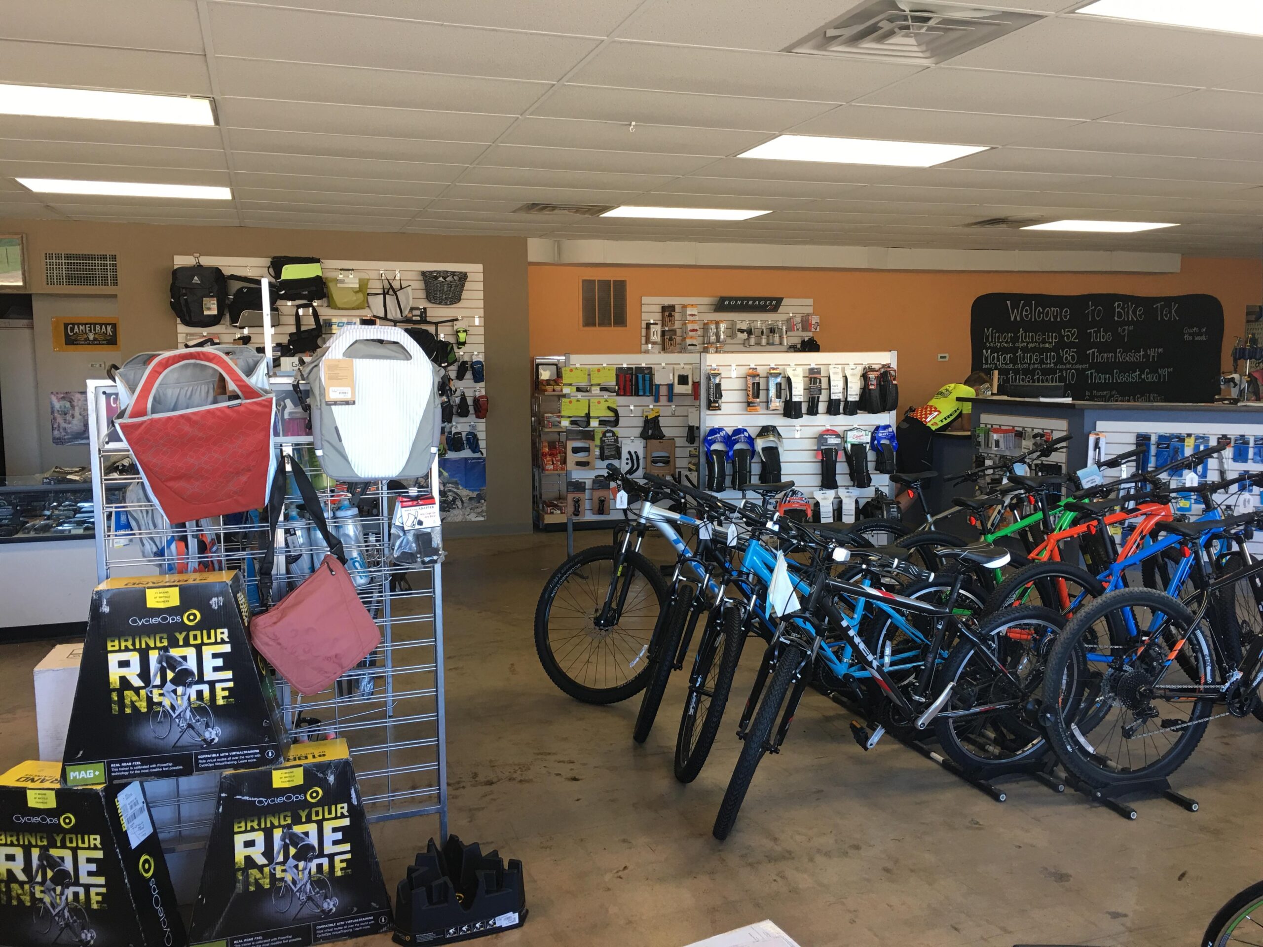 Interior view of a bike shop displaying various bicycles and cycling accessories. In the foreground, there are racks with bicycle bags, and boxes for indoor bike trainers. The background features shelves stocked with bike gear and a chalkboard menu listing services and prices. The shop has a bright and organized layout, inviting cycling enthusiasts.