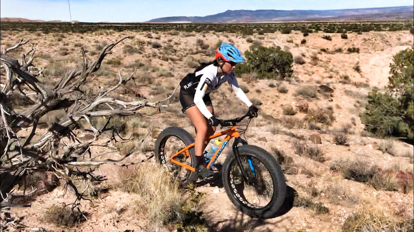 A cyclist riding a fat bike on a rocky, desert terrain, surrounded by sparse vegetation and distant mountains under a clear blue sky. The cyclist is wearing a helmet and a cycling jersey, showcasing an active and adventurous scene. Mariposa Fat Bike Trails mountain bike trail.