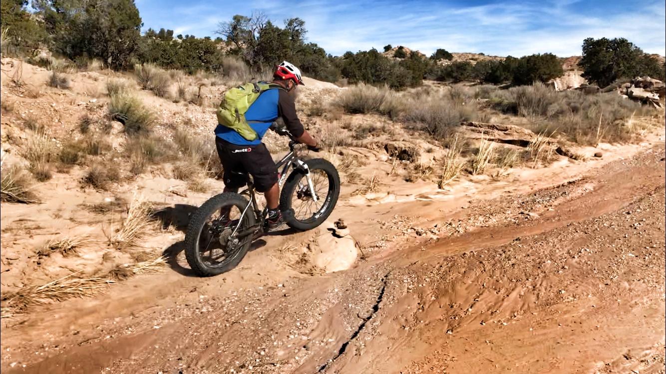 A mountain biker riding on a sandy trail in a desert landscape, surrounded by sparse vegetation and rocky terrain under a clear blue sky. The cyclist is wearing a helmet and a backpack, navigating a path with a slight incline. Mariposa Fat Bike Trails mountain bike trail.
