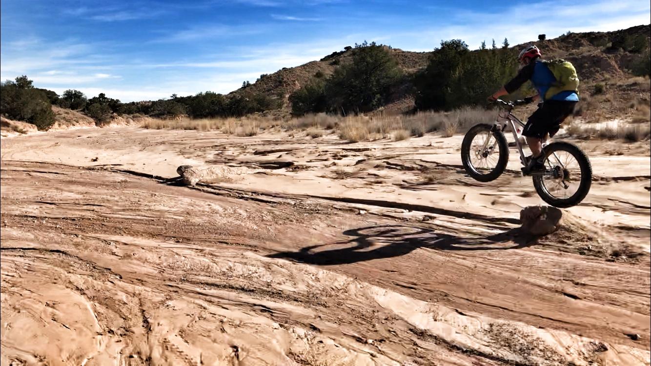 A person riding a fat bike is captured in mid-air as they jump over a rock on a sandy, rugged terrain. In the background, there are dry grasses and sparse vegetation, with hills and a blue sky visible above. Mariposa Fat Bike Trails mountain bike trail.
