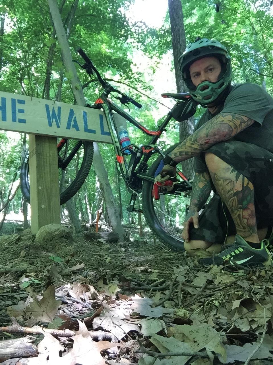 Giant Stance 27.5 2: A person crouches beside a mountain bike in a wooded area, next to a wooden sign that reads "THE WALL." The individual is wearing a helmet and has visible tattoos on their arms, dressed in a gray shirt and camouflage shorts. The ground is covered with leaves and twigs, with trees and greenery in the background.