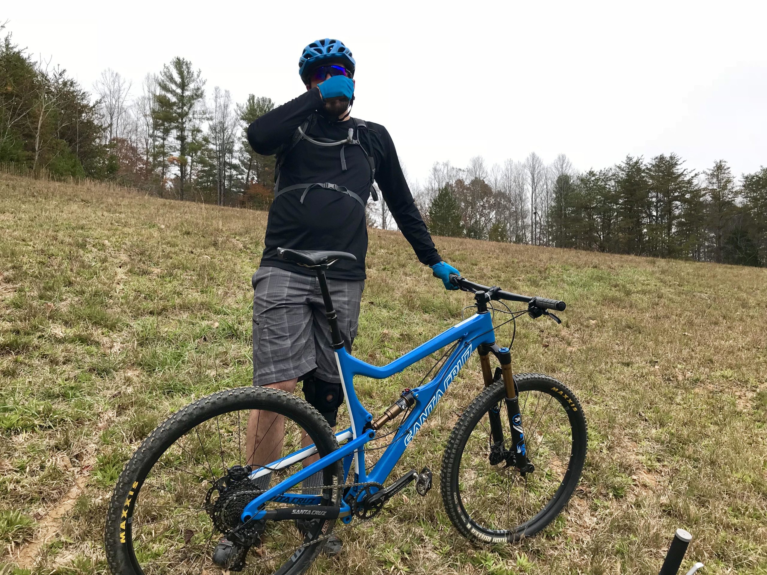 A mountain biker wearing a blue helmet and gloves stands next to a blue Santa Cruz mountain bike in a grassy field. The biker is adjusting a face mask, with trees in the background and a cloudy sky above. The terrain appears rugged, indicating a natural outdoor setting for biking. Warrior Creek mountain bike trail.