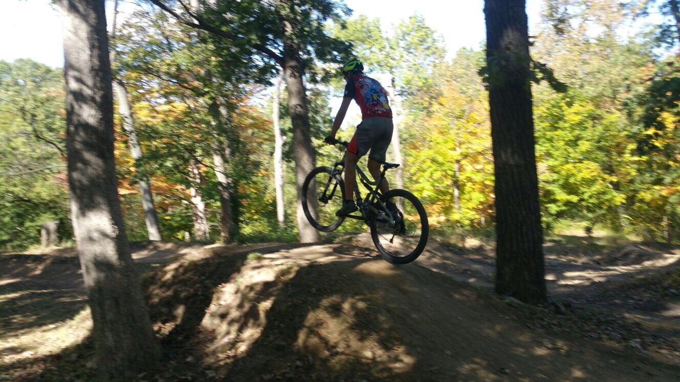 Diamondback Recoil 29: A mountain biker in a colorful jersey is mid-air, jumping off a dirt ramp in a wooded area with vibrant autumn foliage in the background.