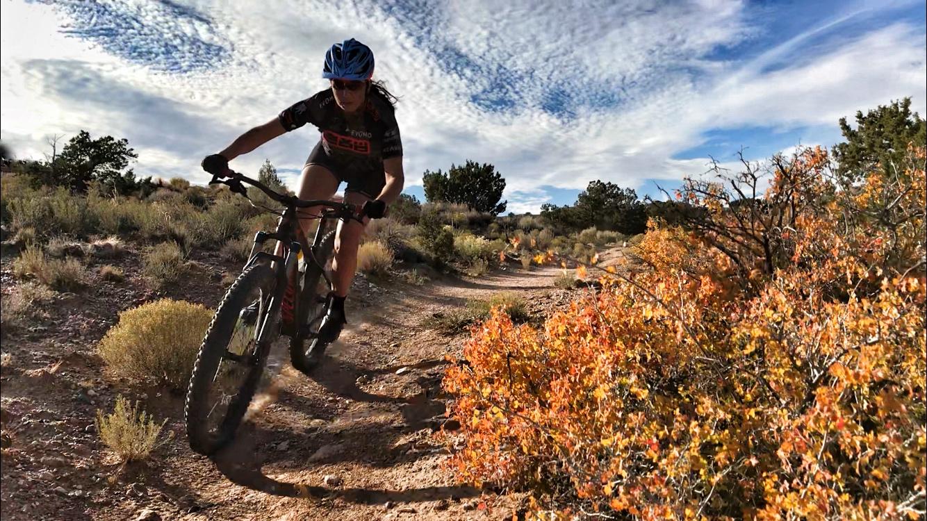 A person riding a mountain bike along a dirt trail surrounded by colorful fall foliage and rugged terrain, under a partly cloudy sky. The rider is leaning forward, demonstrating speed and agility while navigating the trail. Antlion Fatbike trail mountain bike trail.