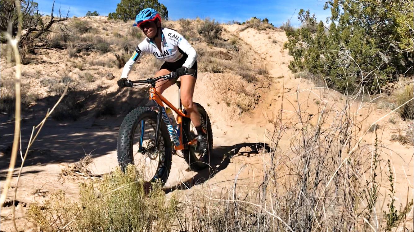 A person riding a fat bike on a sandy trail, surrounded by shrubs and sparse vegetation. They are wearing a blue helmet and a white cycling jersey with colorful accents. The background features a clear blue sky and a rugged landscape. Mariposa Fat Bike Trails mountain bike trail.