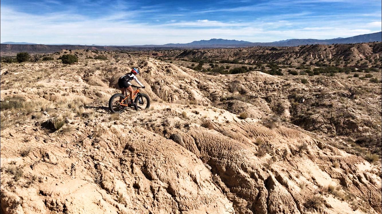 A mountain biker rides on rocky terrain with a vast, desert landscape in the background, featuring rolling hills and blue skies. The biker is dressed in cycling gear and riding a fat bike, highlighting the adventurous spirit of outdoor cycling in rugged conditions. Mariposa Fat Bike Trails mountain bike trail.