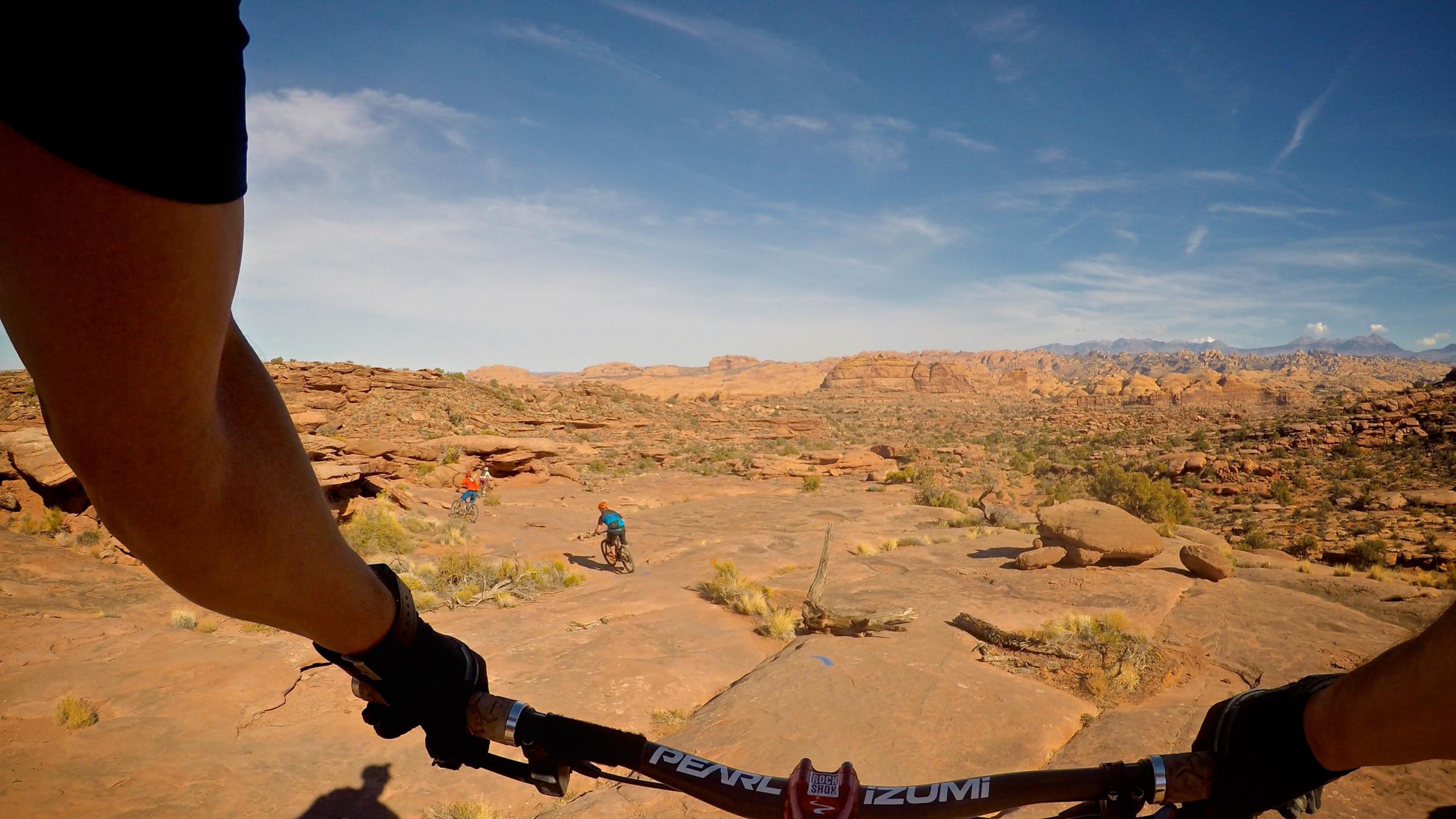 A mountain biker's view from the handlebars, showcasing a rocky landscape with two other cyclists in the background. The scene features arid terrain with sparse vegetation under a clear blue sky. Captain Ahab mountain bike trail.