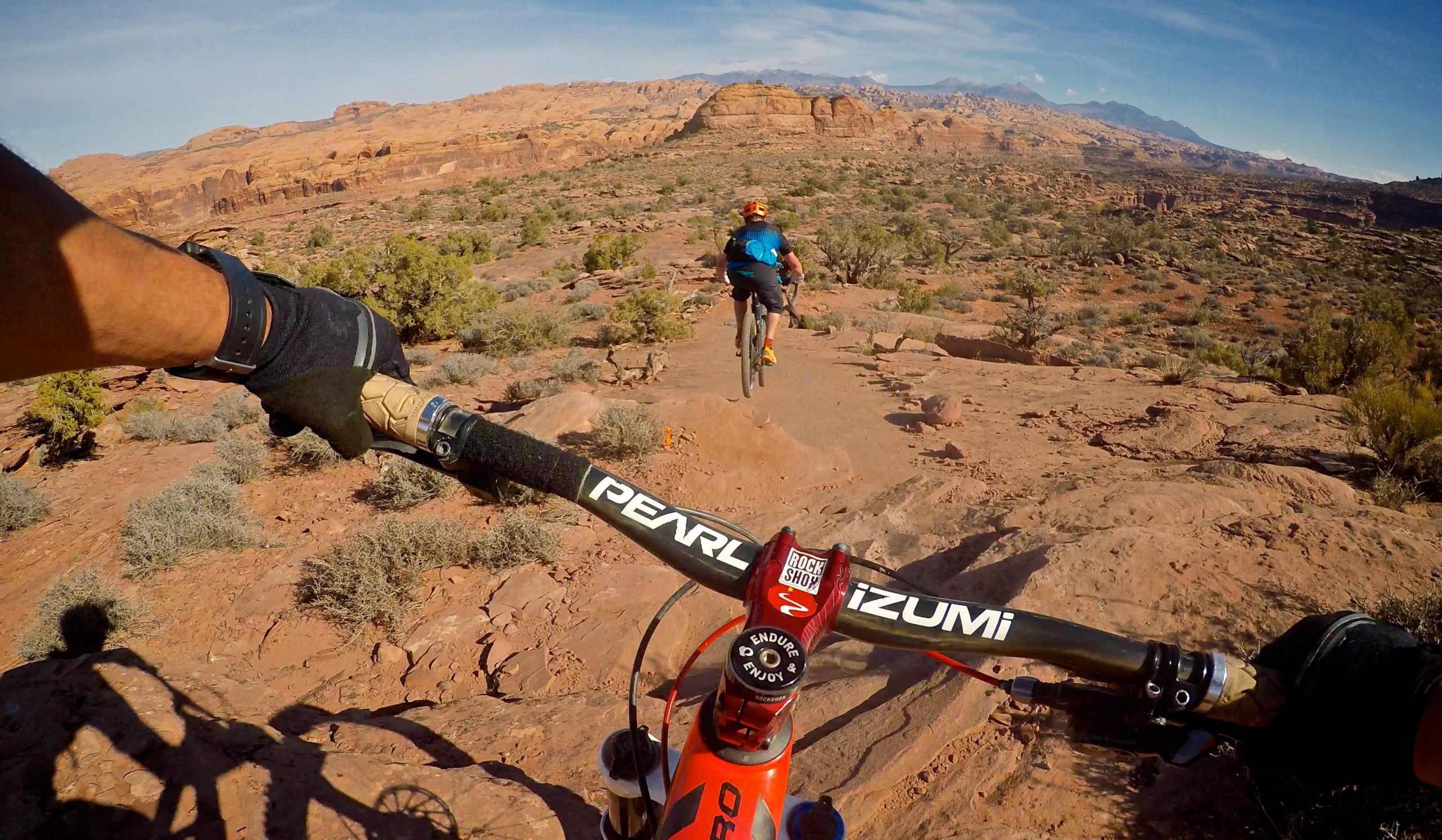 A mountain biker navigates a rocky trail in a desert landscape, with cliffs and shrubs in the background. The perspective shows the rider’s handlebars and a shadow on the ground, while another biker jumps ahead on the trail. Bright blue skies are visible above, highlighting the outdoor adventure setting. Captain Ahab mountain bike trail.