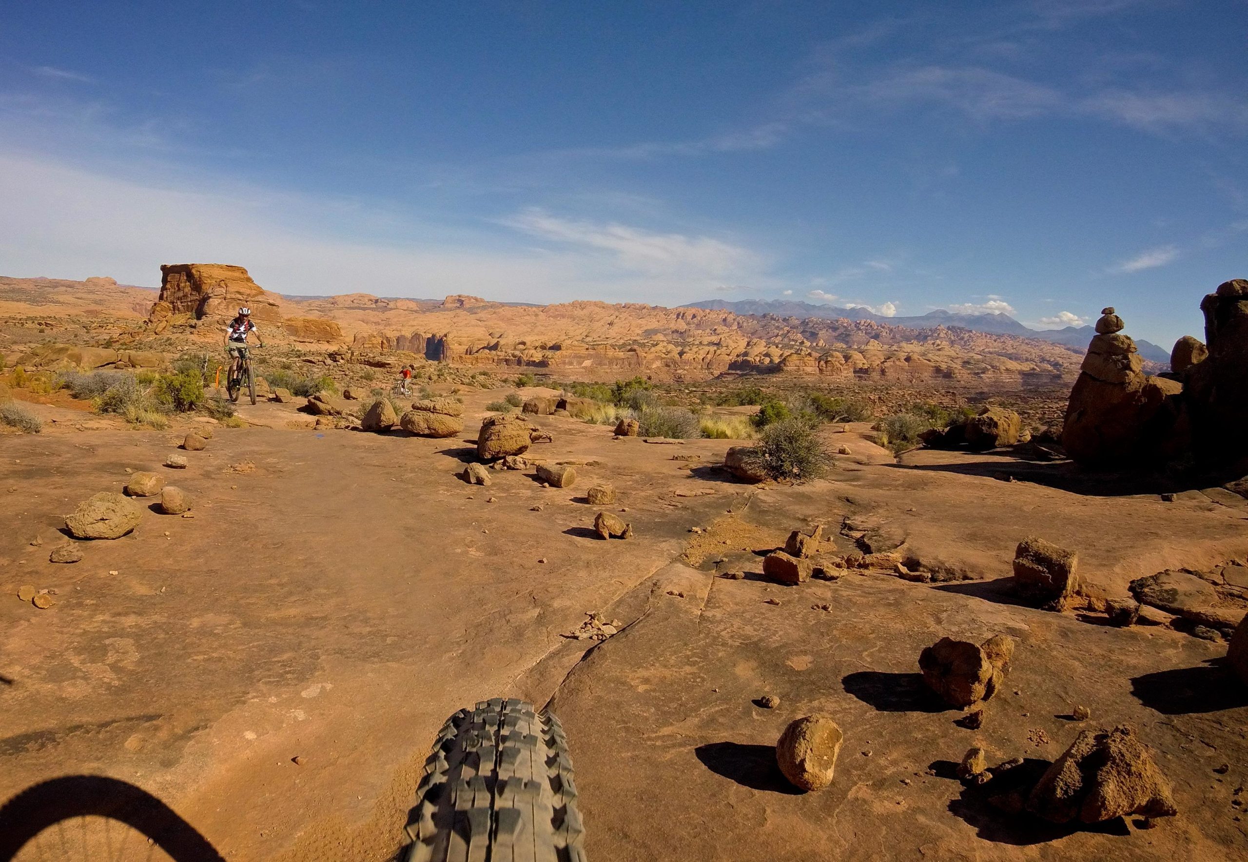 A mountain biker navigating a rocky trail in a desert landscape, surrounded by reddish-brown rock formations and sparse vegetation, under a bright blue sky. Captain Ahab mountain bike trail.