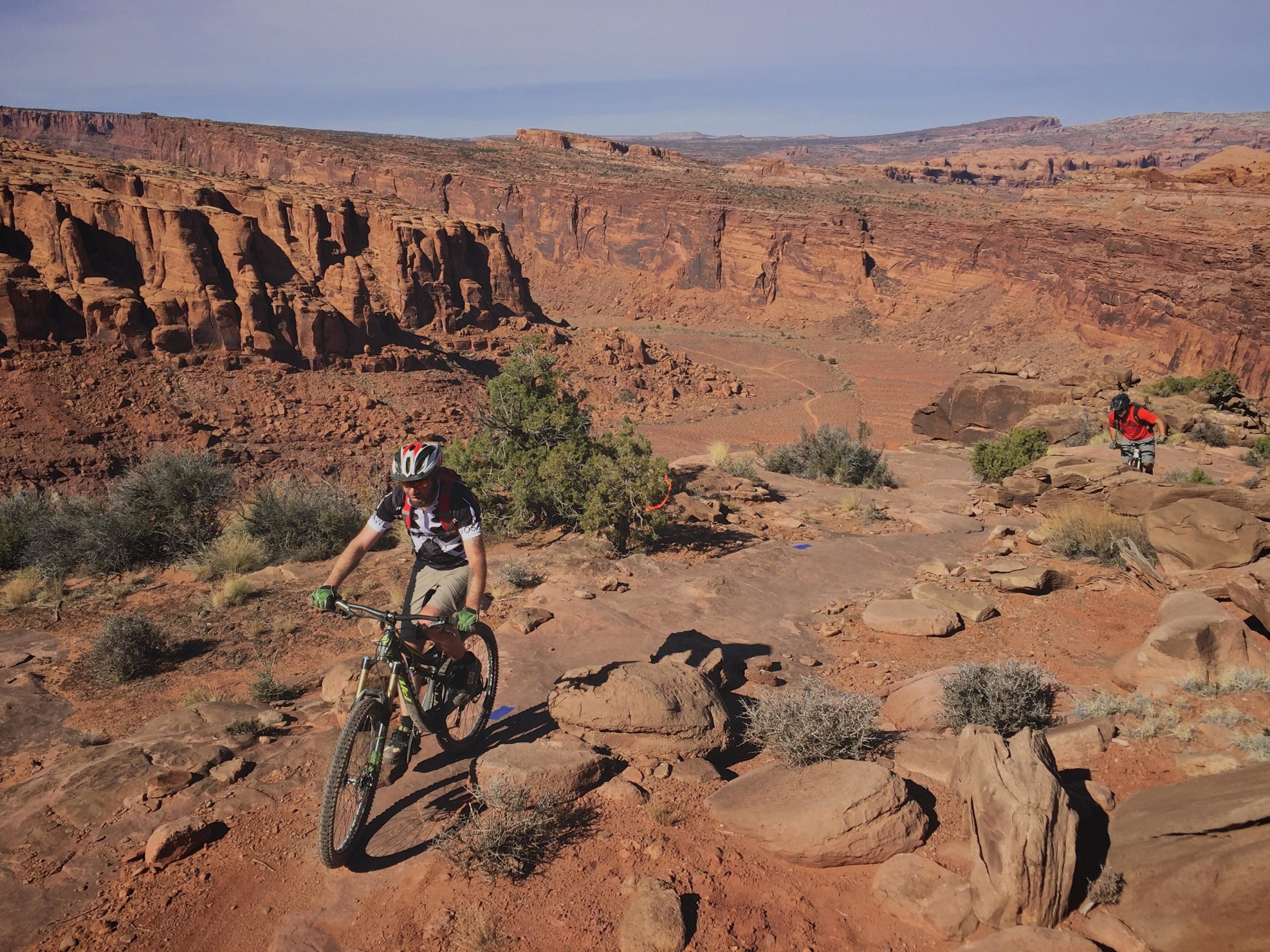 A mountain biker navigating a rocky trail in a desert landscape, surrounded by red cliffs and sparse vegetation. In the background, another person can be seen exploring the area. The sky is clear, indicating a sunny day with a vast and open vista. Captain Ahab mountain bike trail.