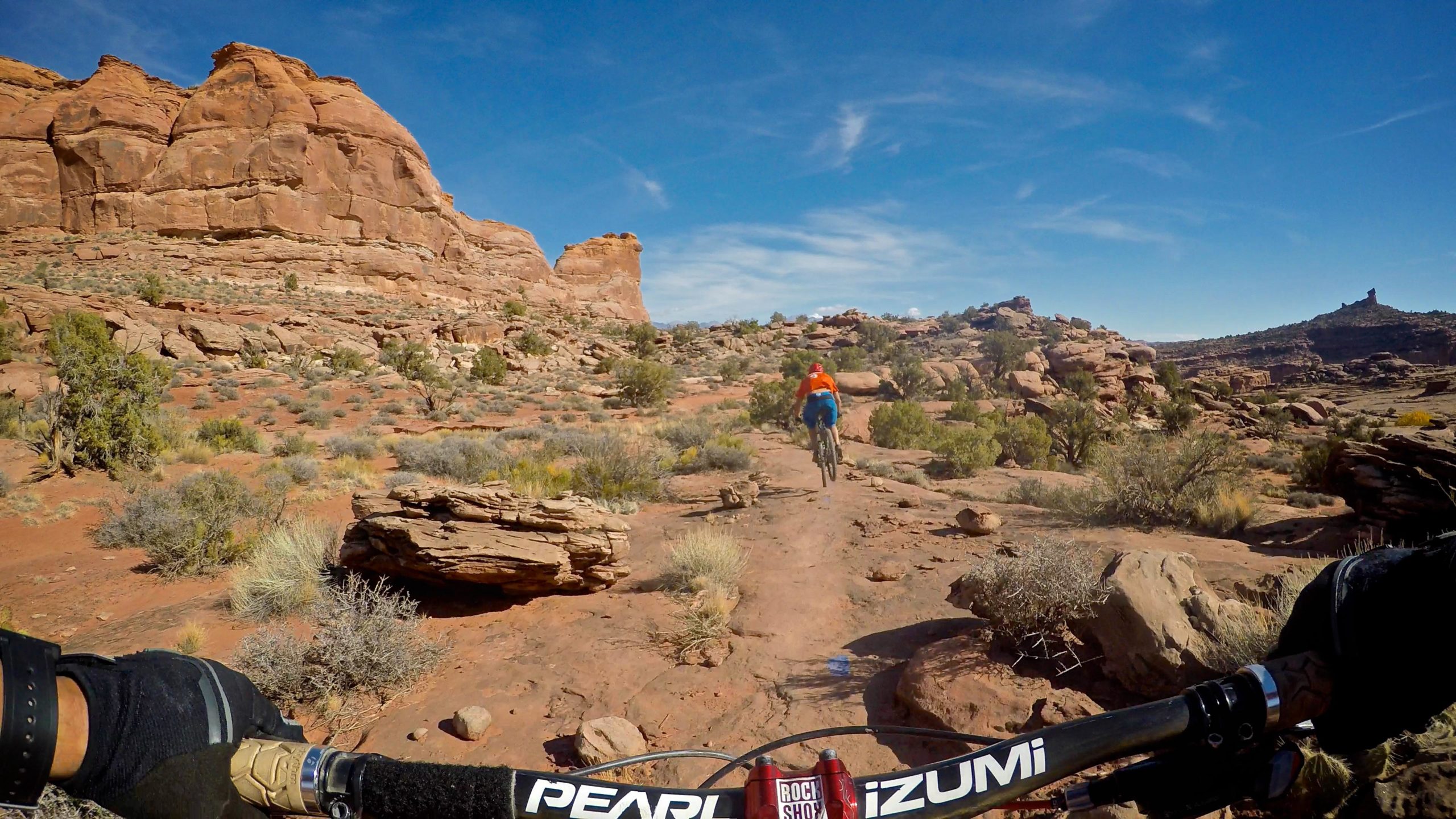 A mountain biker in an orange jacket rides along a dirt trail in a desert landscape, surrounded by rugged red rock formations and sparse vegetation under a clear blue sky. The handlebars of the bike are visible in the foreground, emphasizing the biking perspective. Captain Ahab mountain bike trail.