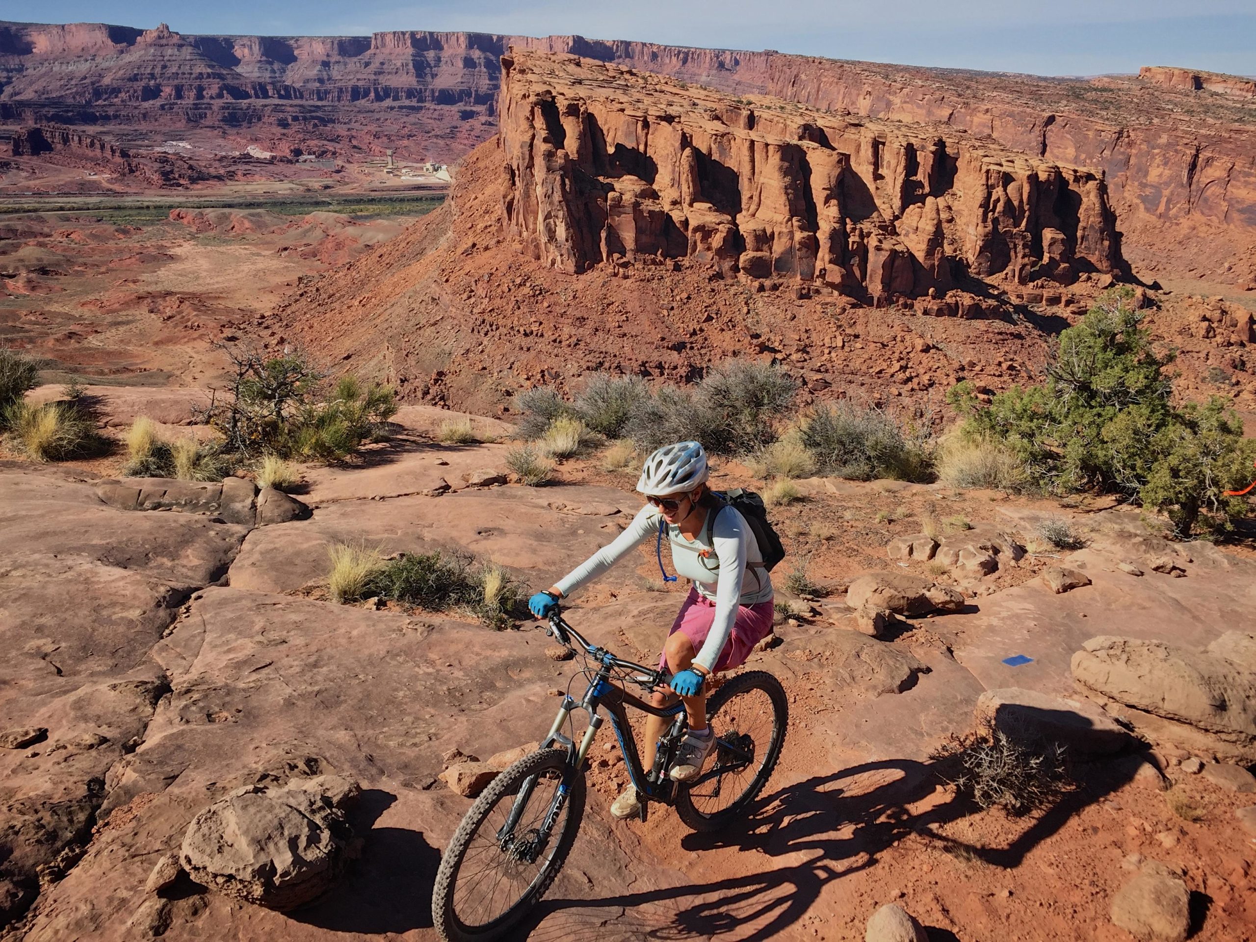 A mountain biker navigating a rocky trail in a desert landscape, with red rock formations and a clear blue sky in the background. The biker is wearing a helmet, gloves, and a backpack, focusing on the challenging terrain. Captain Ahab mountain bike trail.