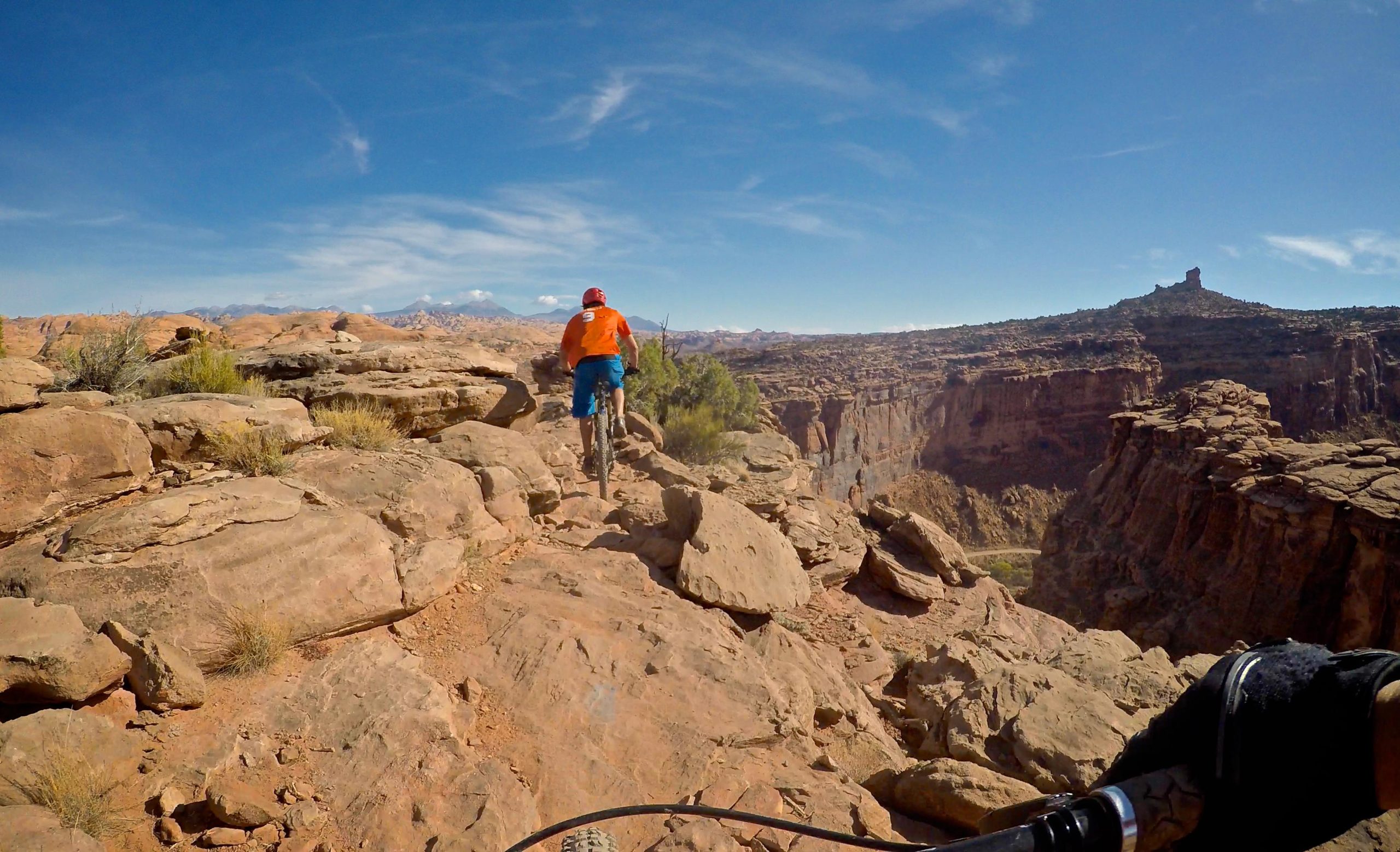 A mountain biker in an orange shirt and blue shorts navigates a rocky trail overlooking a canyon, with blue skies and distant mountains in the background. Captain Ahab mountain bike trail.
