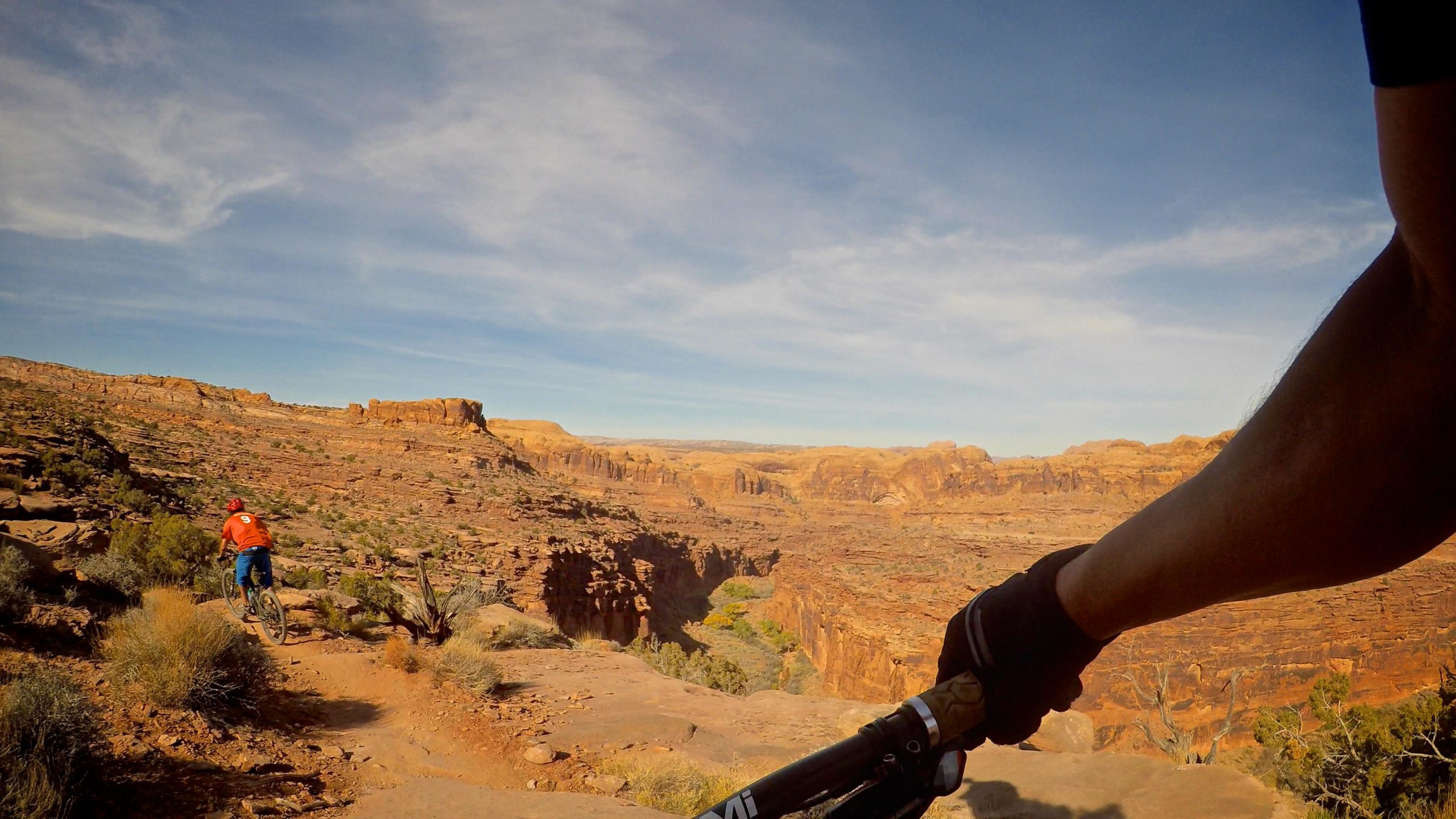 A mountain biker rides along a rocky trail with a canyon landscape in the background, showcasing dramatic cliffs and a clear blue sky. The biker is seen from the perspective of their handlebars, emphasizing the rugged terrain and scenic views. Captain Ahab mountain bike trail.