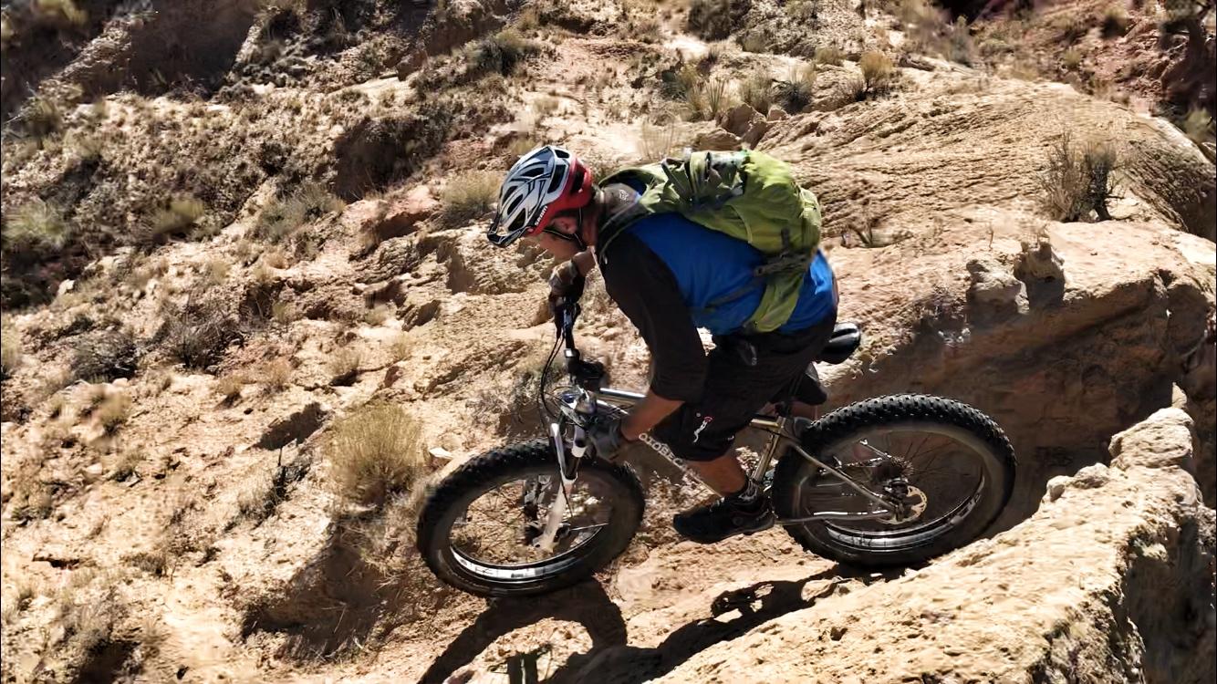 A mountain biker navigating a rocky trail, wearing a helmet and a green backpack, as they descend a steep, uneven surface. The surrounding terrain features dry brush and rocky outcrops typical of a rugged outdoor landscape. Mariposa Fat Bike Trails mountain bike trail.