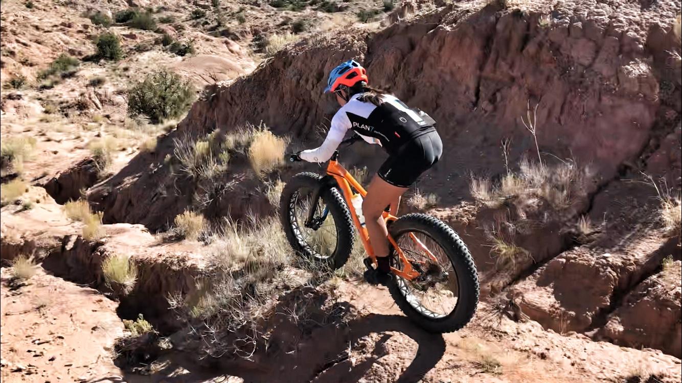 A person riding a fat bike down a rocky slope in a dry, desert landscape, with sparse vegetation and steep terrain visible in the background. The rider is wearing a helmet and athletic gear, demonstrating a dynamic biking maneuver. Mariposa Fat Bike Trails mountain bike trail.